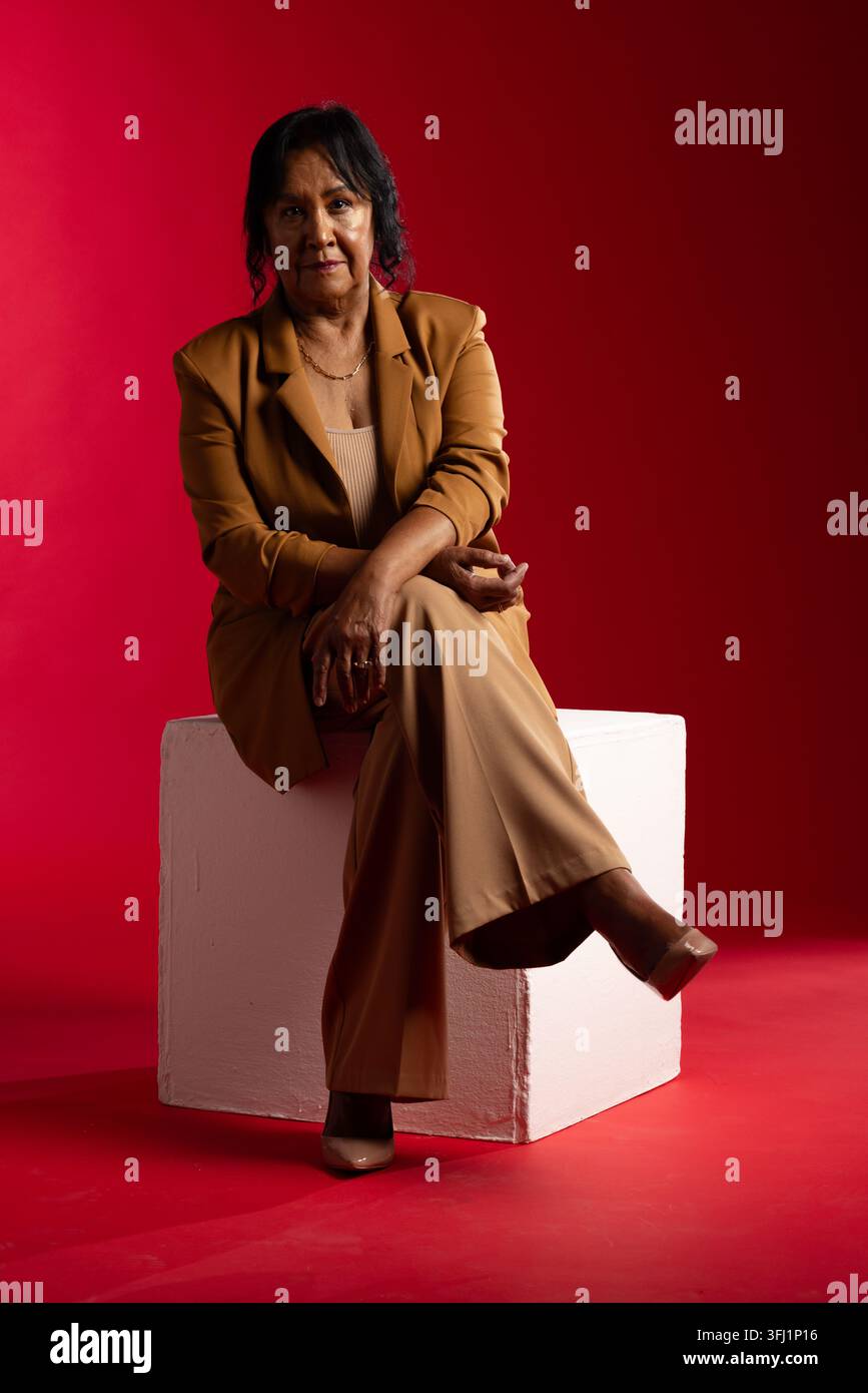 Donna anziana africana seduta su un cubo bianco in studio, indossa un blazer marrone chiaro e pantaloni a gamba larga Foto Stock