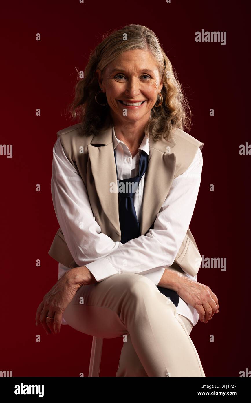 Donna anziana seduta su uno sgabello indossa una camicia bianca, un gilet beige e una cravatta blu navy, sorridente Foto Stock