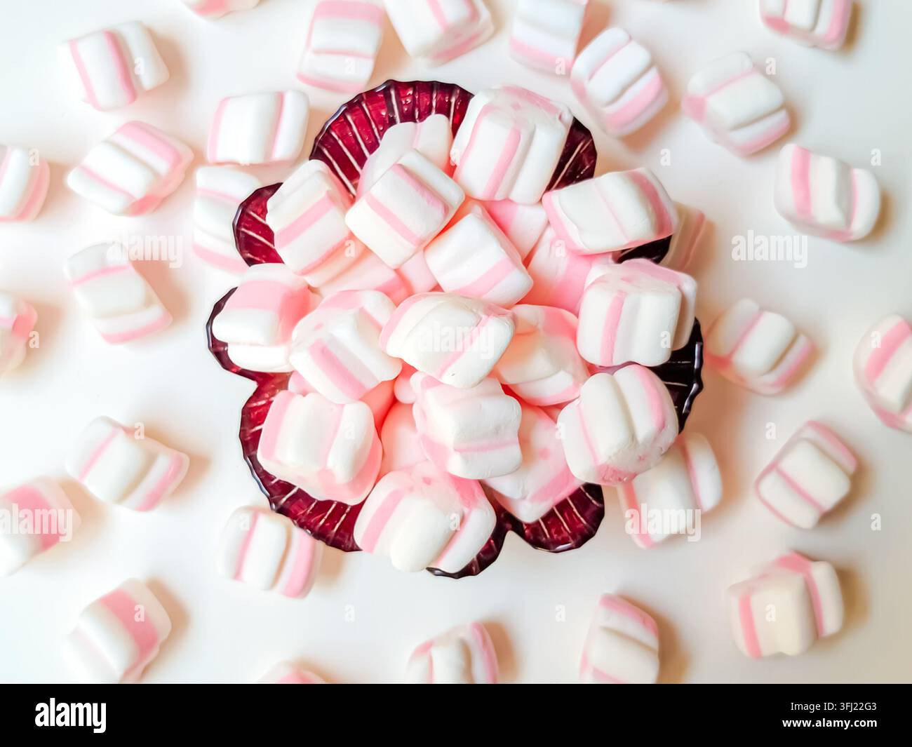Vista dall'alto di mini marshmallow rosa e bianchi a strisce in una ciotola e sparsi su uno sfondo bianco. Foto Stock