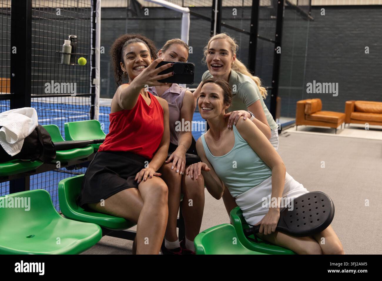 Diverse amiche in posa per un selfie al campo del padel con un abbigliamento sportivo che regge smartphone e racchetta. Compagni di squadra, gruppo, gioia, vibrante, colorato, social, Foto Stock