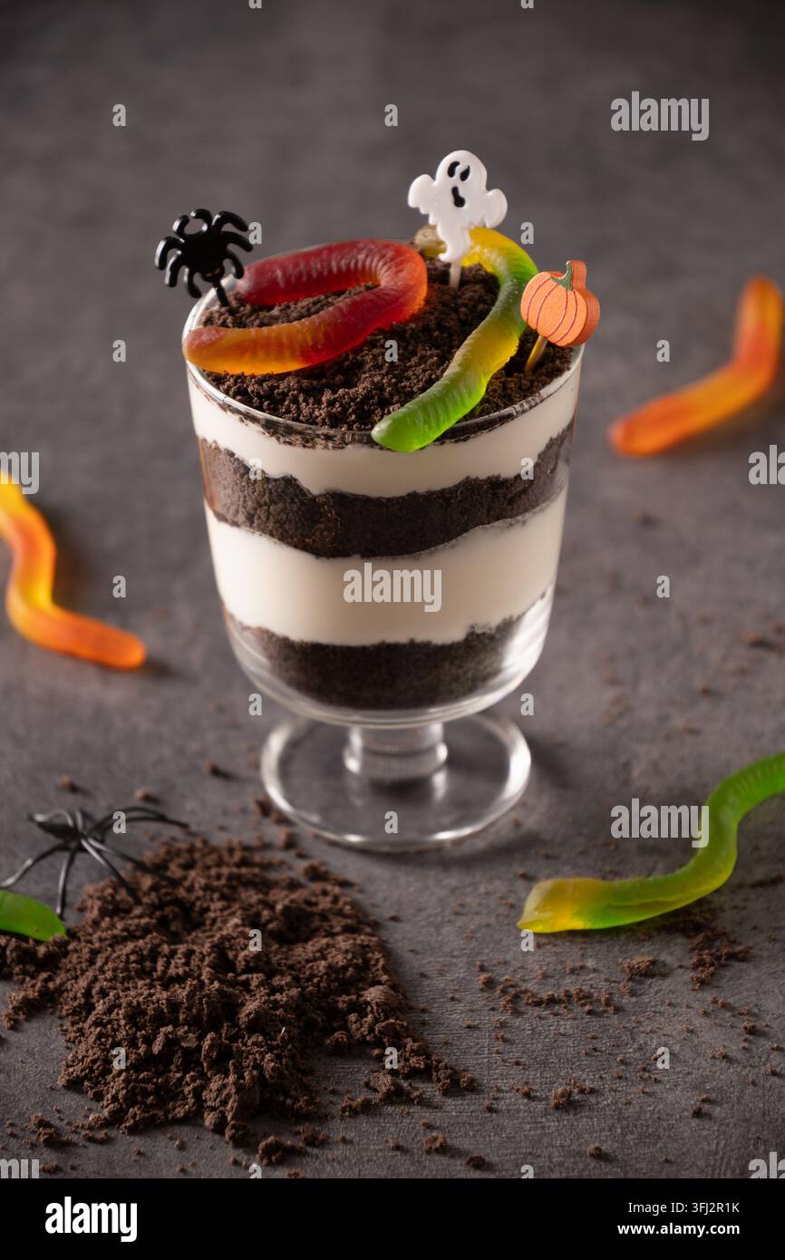 Torta di terra individuale di Halloween a strati con briciole di cioccolato e panna, sormontata da vermi di gelatina, decorazioni a fantasma, ragni e zucca per una festa spaventosa Foto Stock