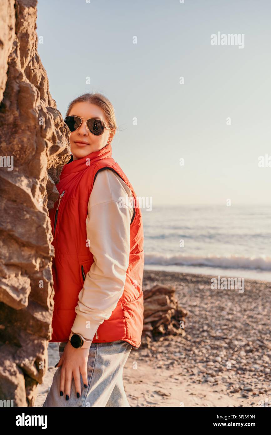 Donna sulla spiaggia, costa, in piedi su una scogliera al tramonto mentre indossa un giubbotto rosso e occhiali da sole alla moda Foto Stock