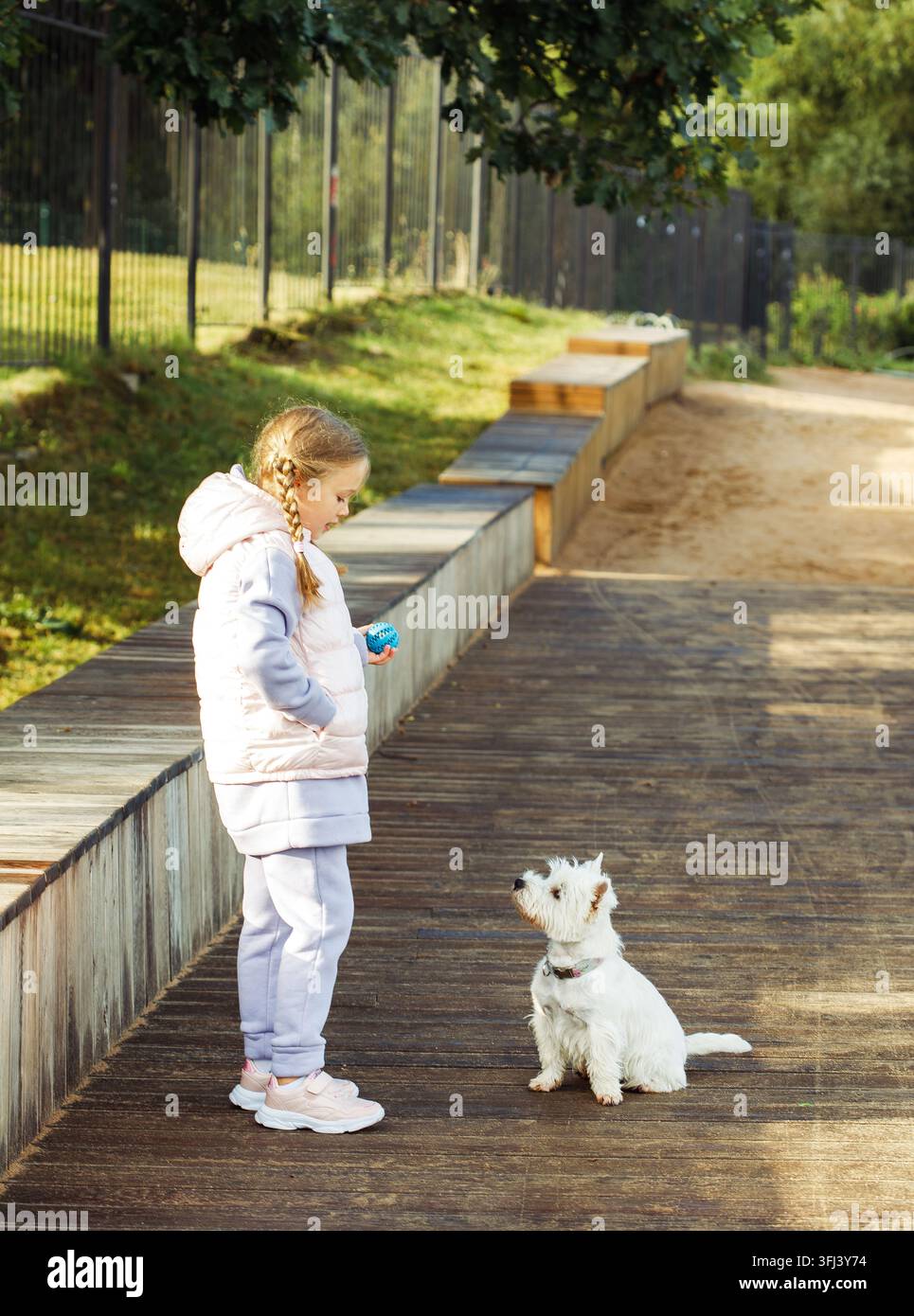 Una ragazza con le trecce gioca con un piccolo cane bianco e sorride felice. Foto Stock