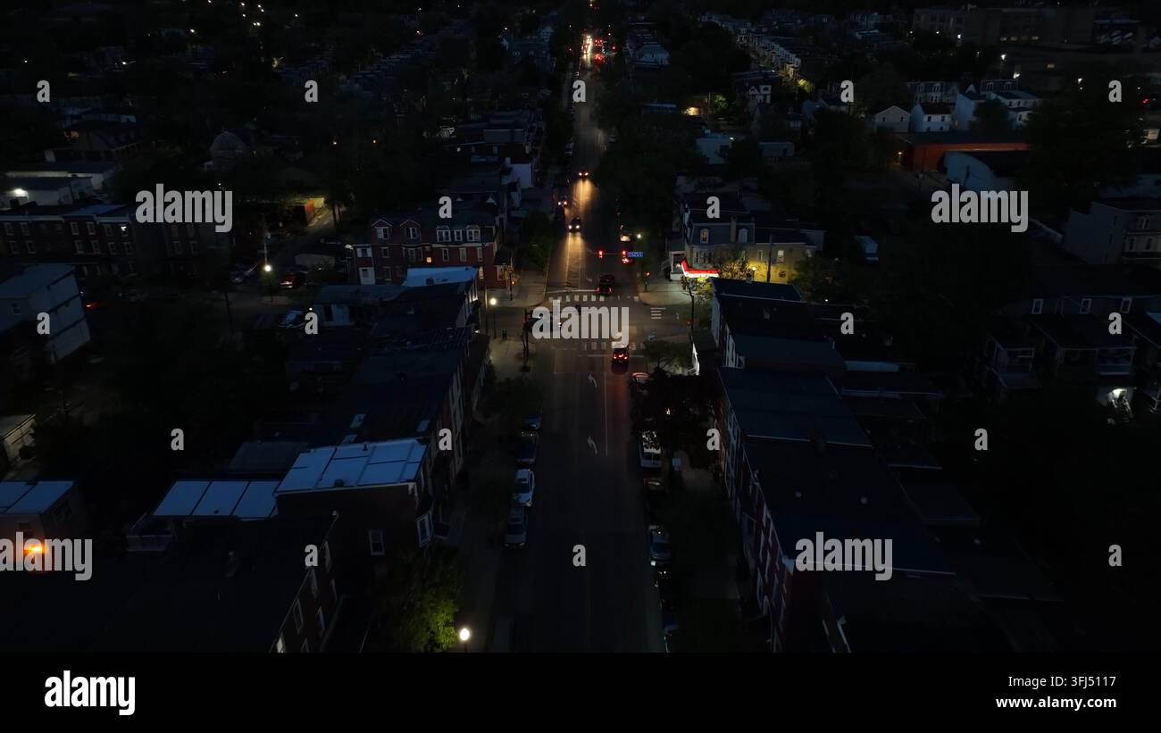 Vista notturna di un incrocio urbano in una città americana. Strade fiancheggiate da case a schiera e lampioni fioca. I fari delle auto creano scie di luce lungo th Foto Stock