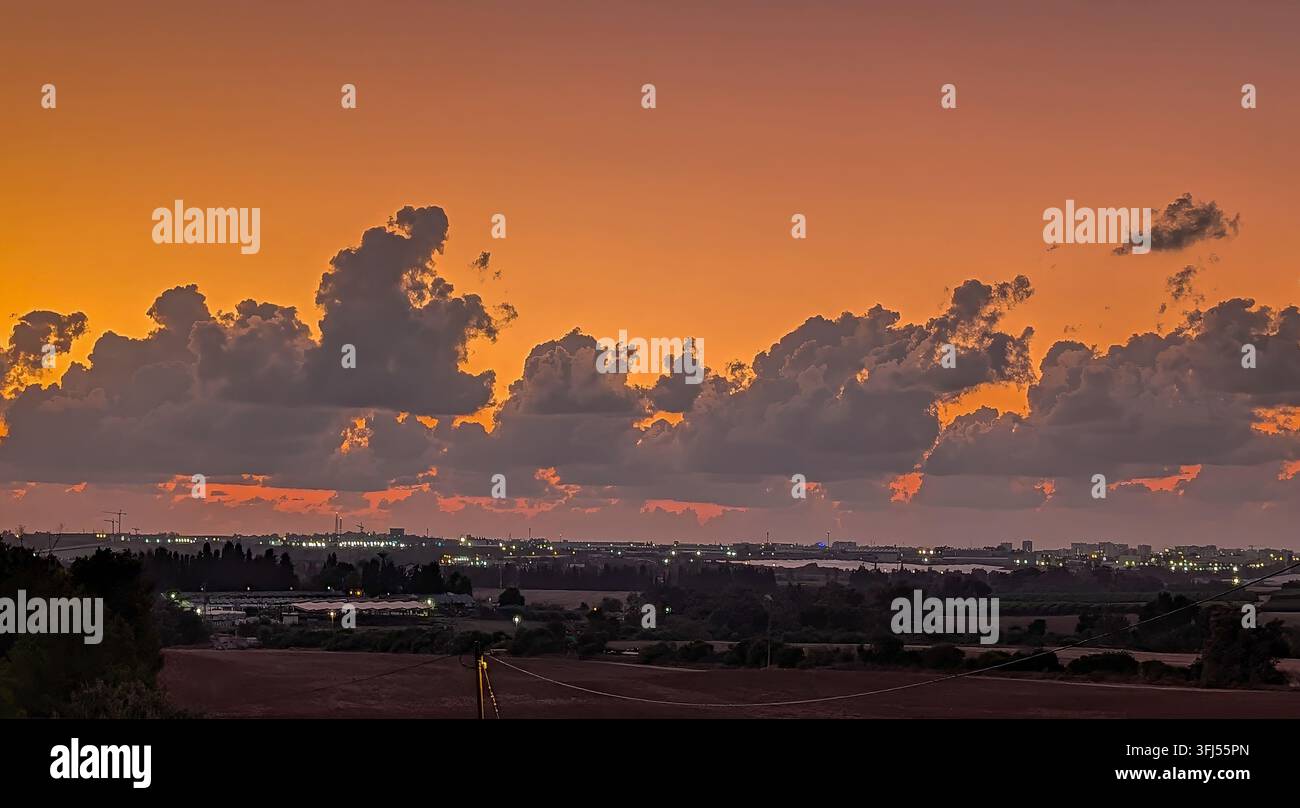 Un tramonto sorprendente e infuocato sulla città di Sderot, Israele, mentre le luci della città si accendono e luccicano sotto le nuvole che nascondono il sole Foto Stock