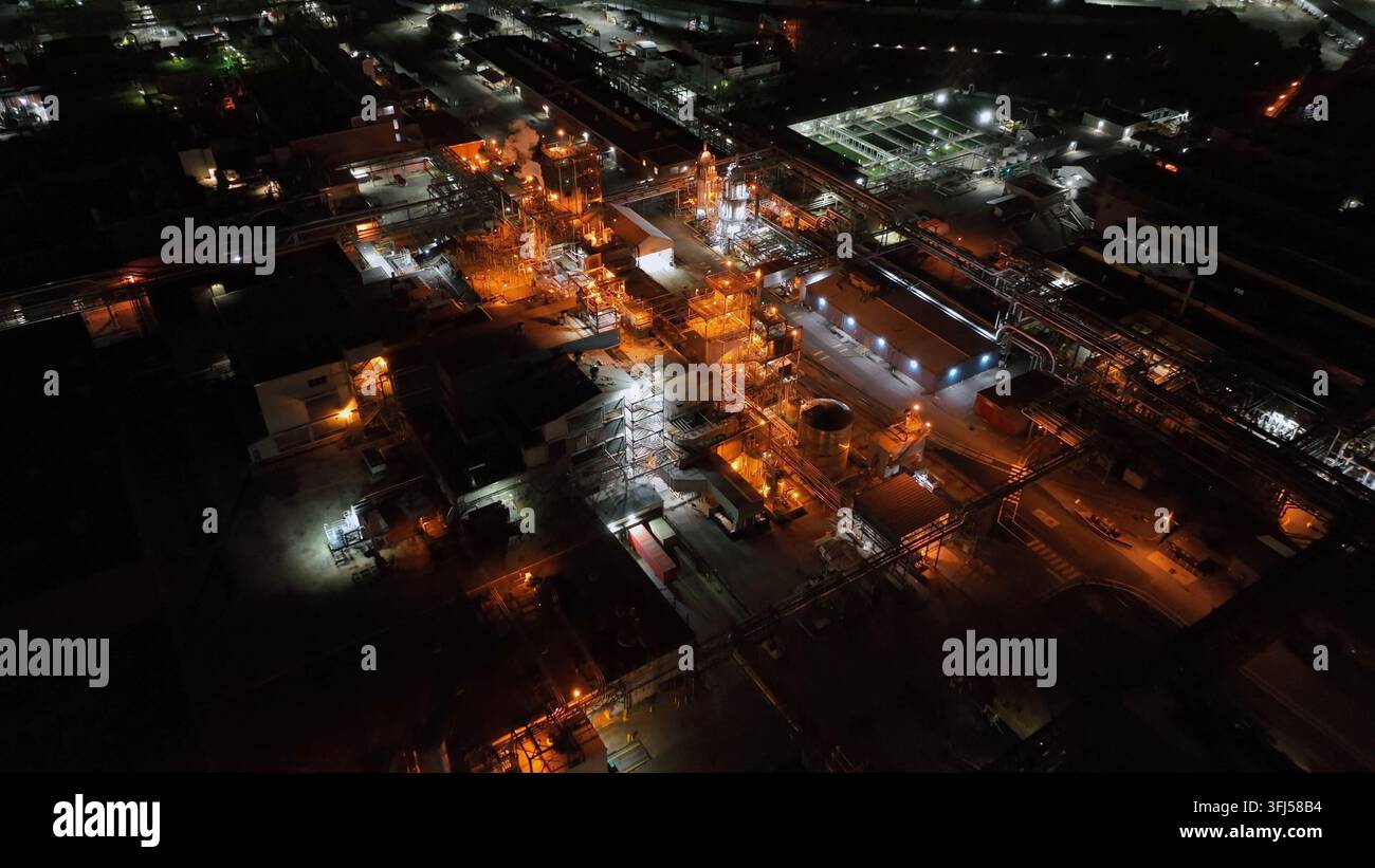 Ampia struttura industriale di notte, che mostra strutture arancioni luminose, vapore e complessi sistemi di tubazioni in un vasto complesso produttivo. Ae Foto Stock