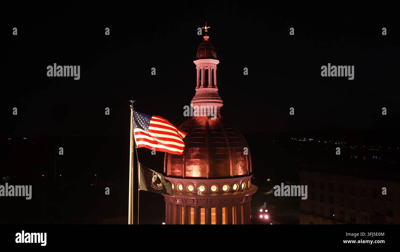 Bandiera americana sventola accanto al cupo di rame illuminato del New Jersey State House a Trenton, New Jersey di notte, una lanterna luminosa e colonne si ergono contro Foto Stock