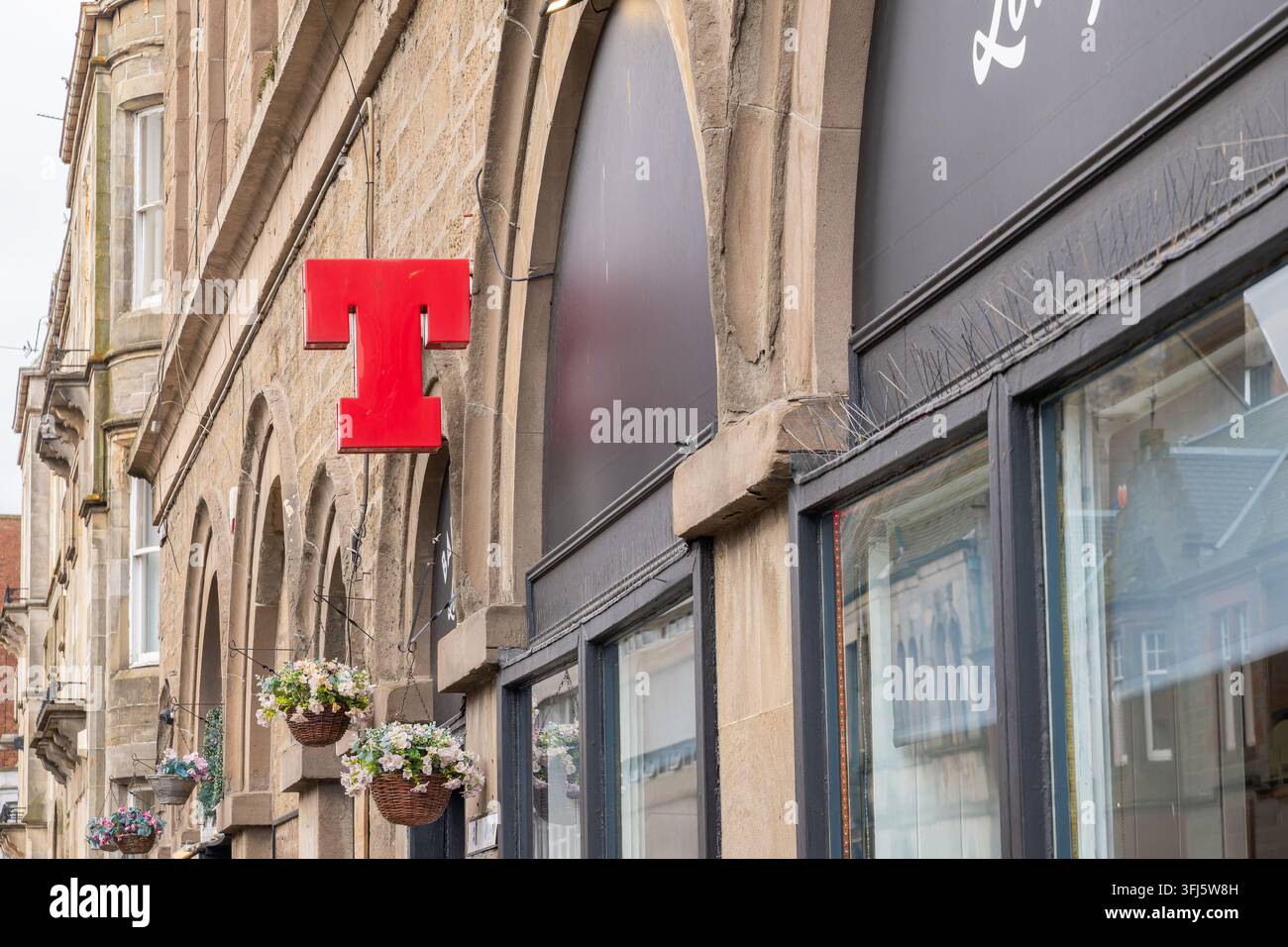 Insegna rossa della birra Tennents con lettera T fissata a un'arco di arenaria vittoriana sopra cesti di fiori sospesi, accanto a un bar con facciata scura e punte anti-uccelli, D Foto Stock