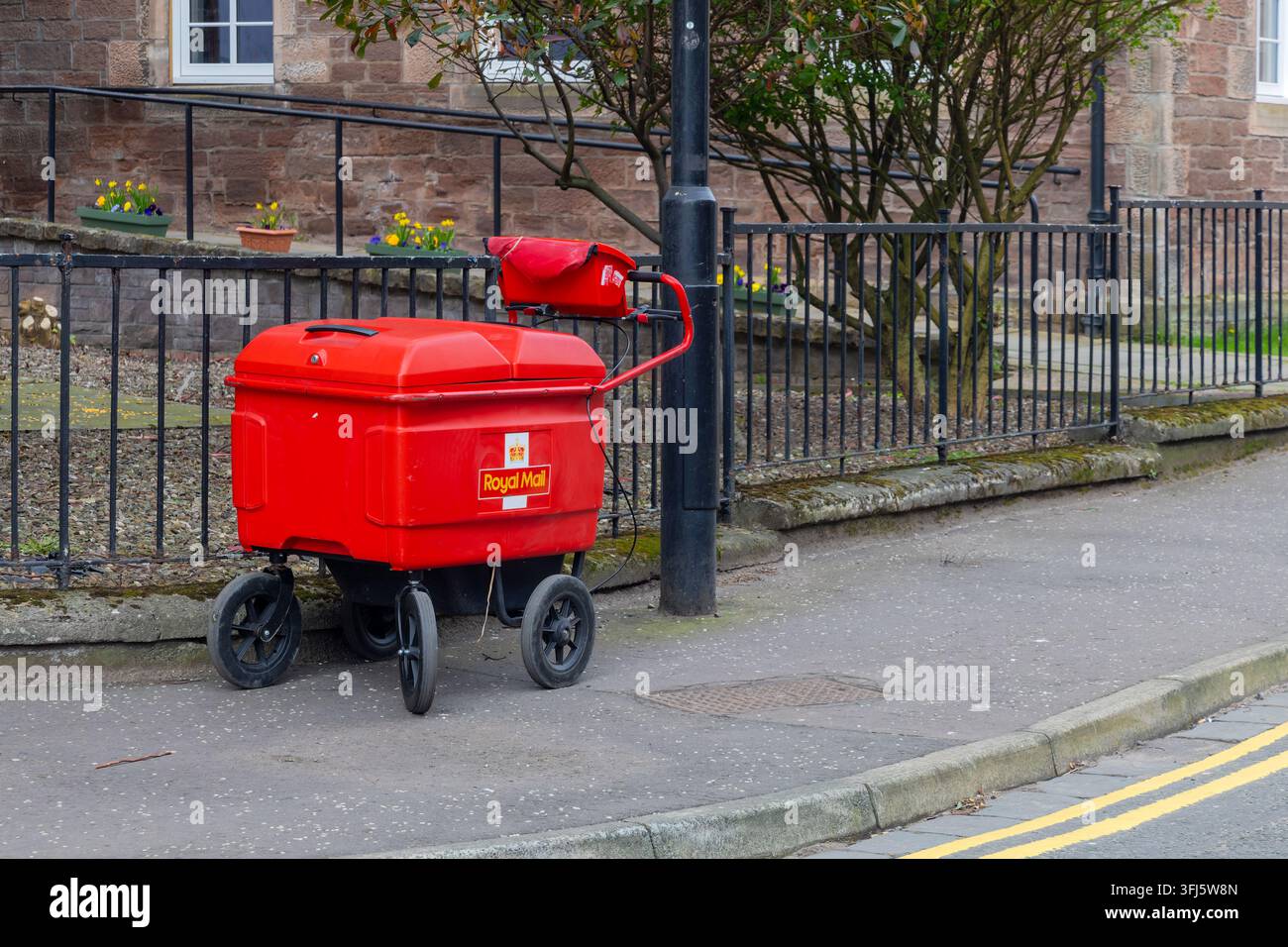 Carrello di consegna a quattro ruote Royal Mail rosso acceso fissato a un lampione in ghisa su un marciapiede residenziale tranquillo, con una borsa postale rossa appoggiata sopra Foto Stock