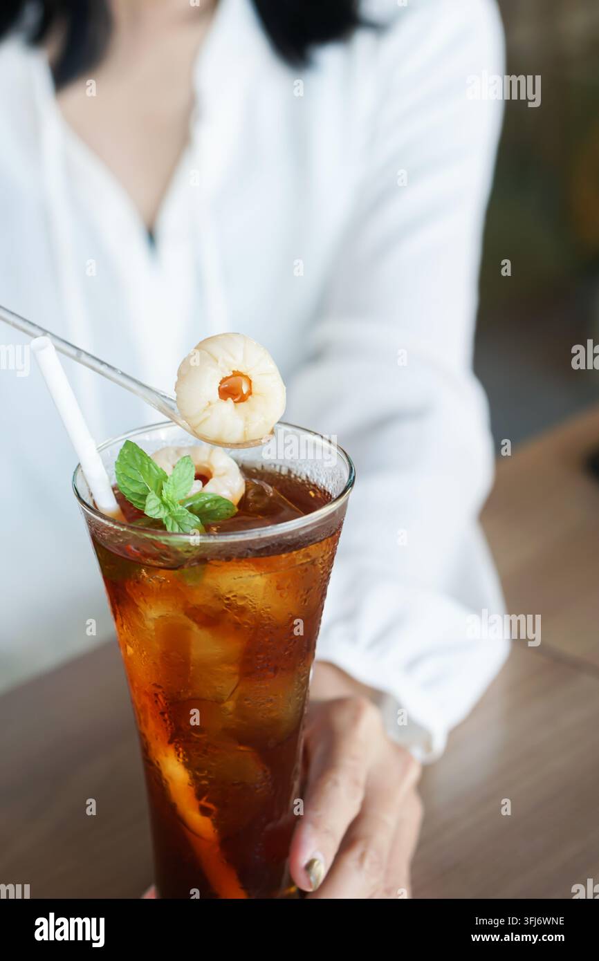 Primo piano di una donna che tiene un alto cocktail di tè freddo con litchi, menta e una cannuccia a un tavolo da caffè. Foto Stock
