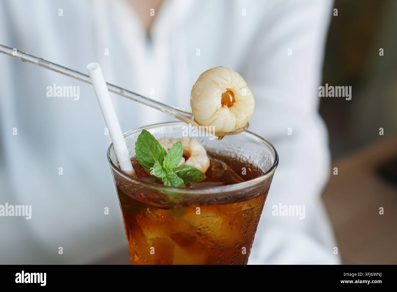 Primo piano di un tè freddo in un bicchiere con frutti di litchi, menta fresca, cubetti di ghiaccio e una cannuccia, tenuto da una persona con una camicia bianca. Foto Stock