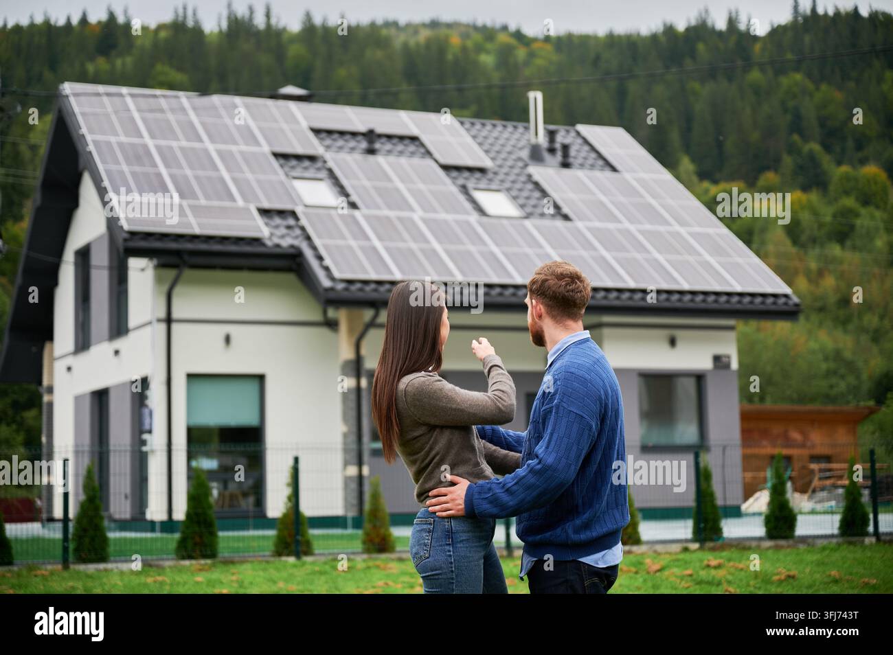 Coppia che osserva una casa con pannelli solari, donna che indica il sistema sul tetto. Proprietari di casa che pianificano l'uso di energie rinnovabili, casa ecologica e concetto di stile di vita sostenibile Foto Stock