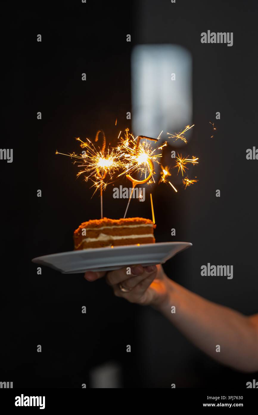Mano che presenta una fetta di torta ripiena di panna sormontata da decorazioni a forma di numeri scintillanti, illustrando la presentazione di cibi per compleanni e l'ospitalità festosa se Foto Stock
