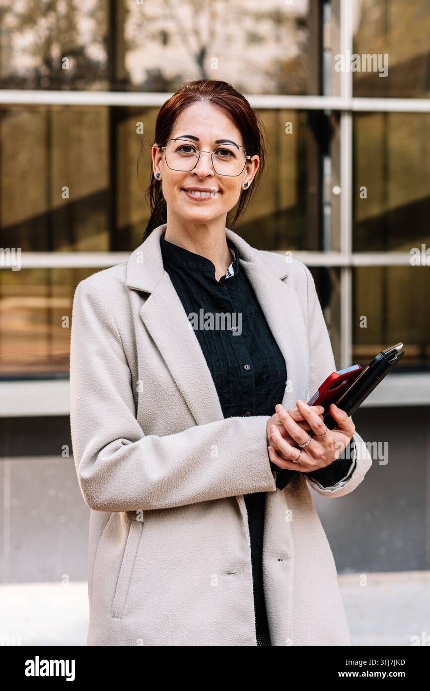 Donna d'affari in piedi davanti a un edificio per uffici con dispositivi in mano Foto Stock