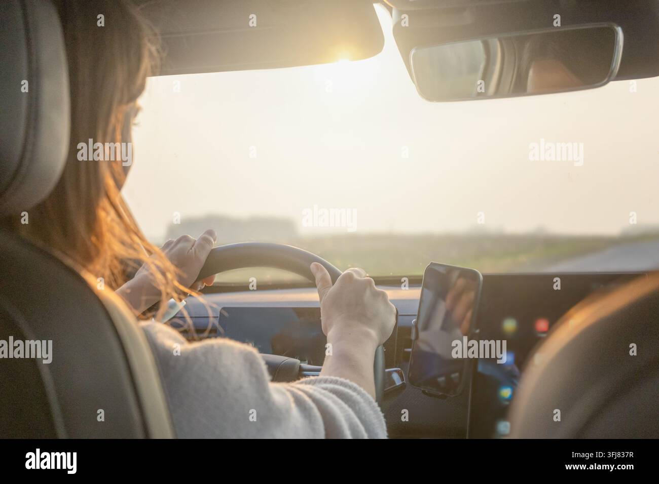 Donna guida un'auto con sterzo a destra nel traffico a sinistra con entrambe le mani sul volante e uno smartphone montato per la navigazione. Guida al tramonto Foto Stock