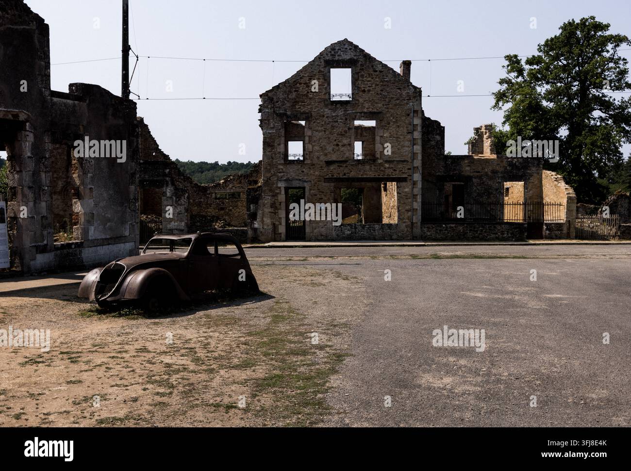Oradour-sur-Glane, Francia. 28 luglio 2026: Scocca di auto arrugginita degli anni '40 davanti alle case in pietra in rovina della piazza principale. Foto Stock