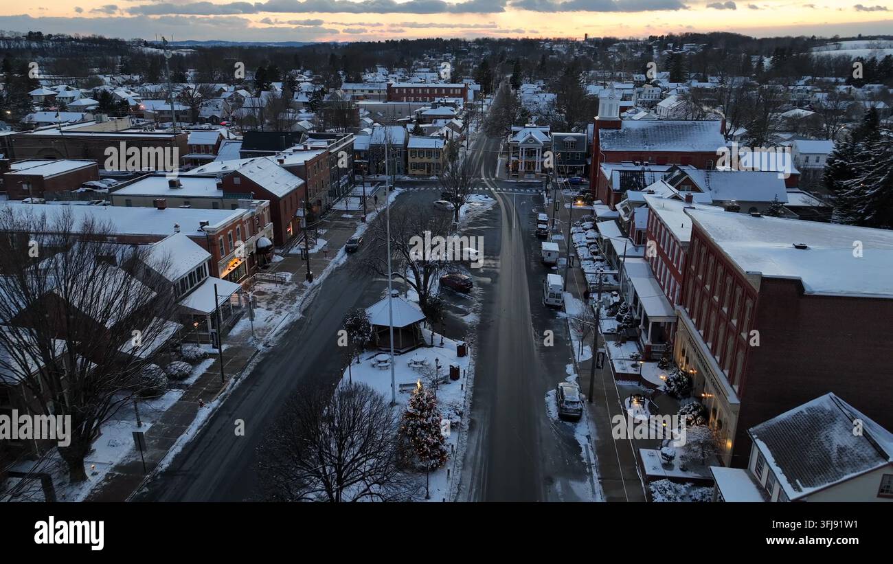 Vista aerea con drone di una piccola città storica al tramonto in inverno. La neve copre i tetti e le strade mentre le auto si muovono lentamente attraverso la città tranquilla, creando Foto Stock