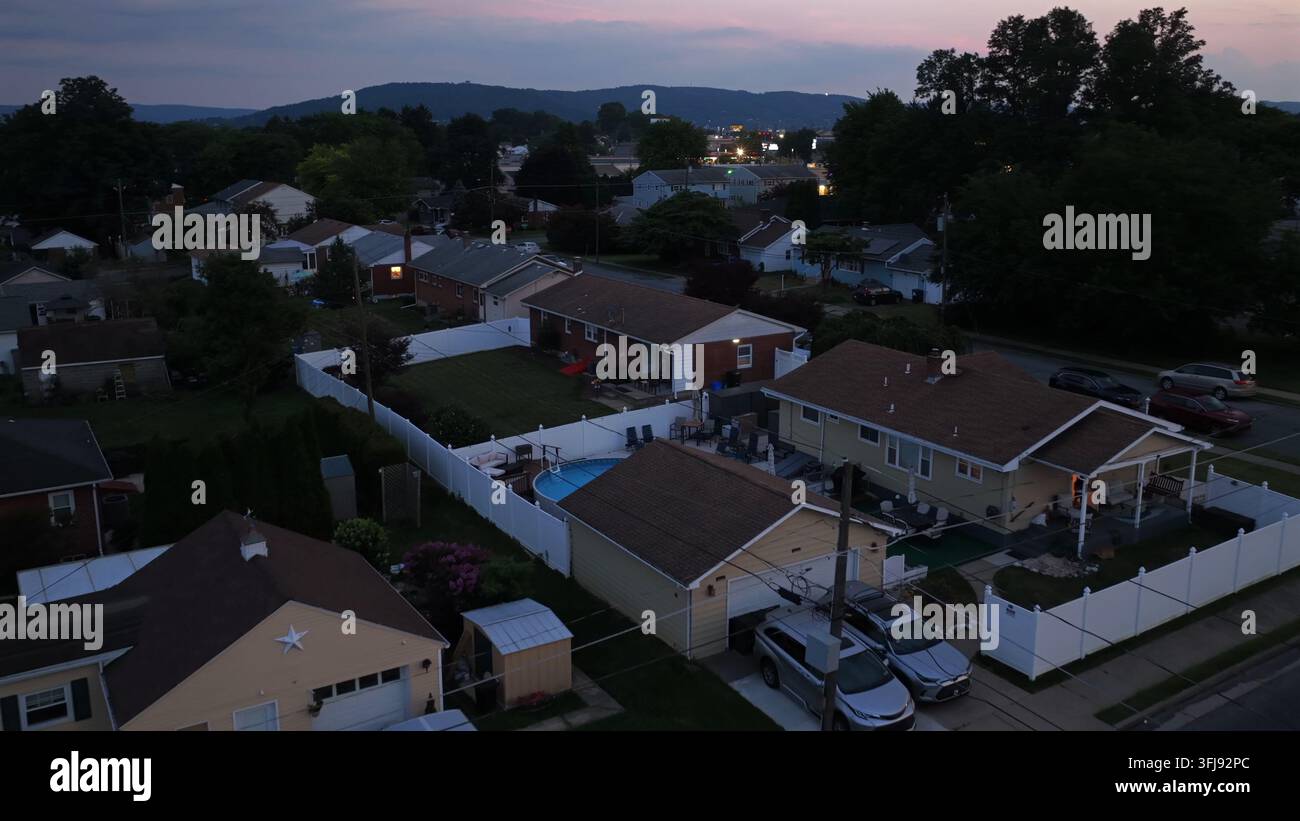 La piscina nel cortile si trova accanto a case a un piano con auto nei vialetti nel Lehigh Valley, Pennsylvania al crepuscolo, le colline lontane e le luci della città brillano oltre Foto Stock