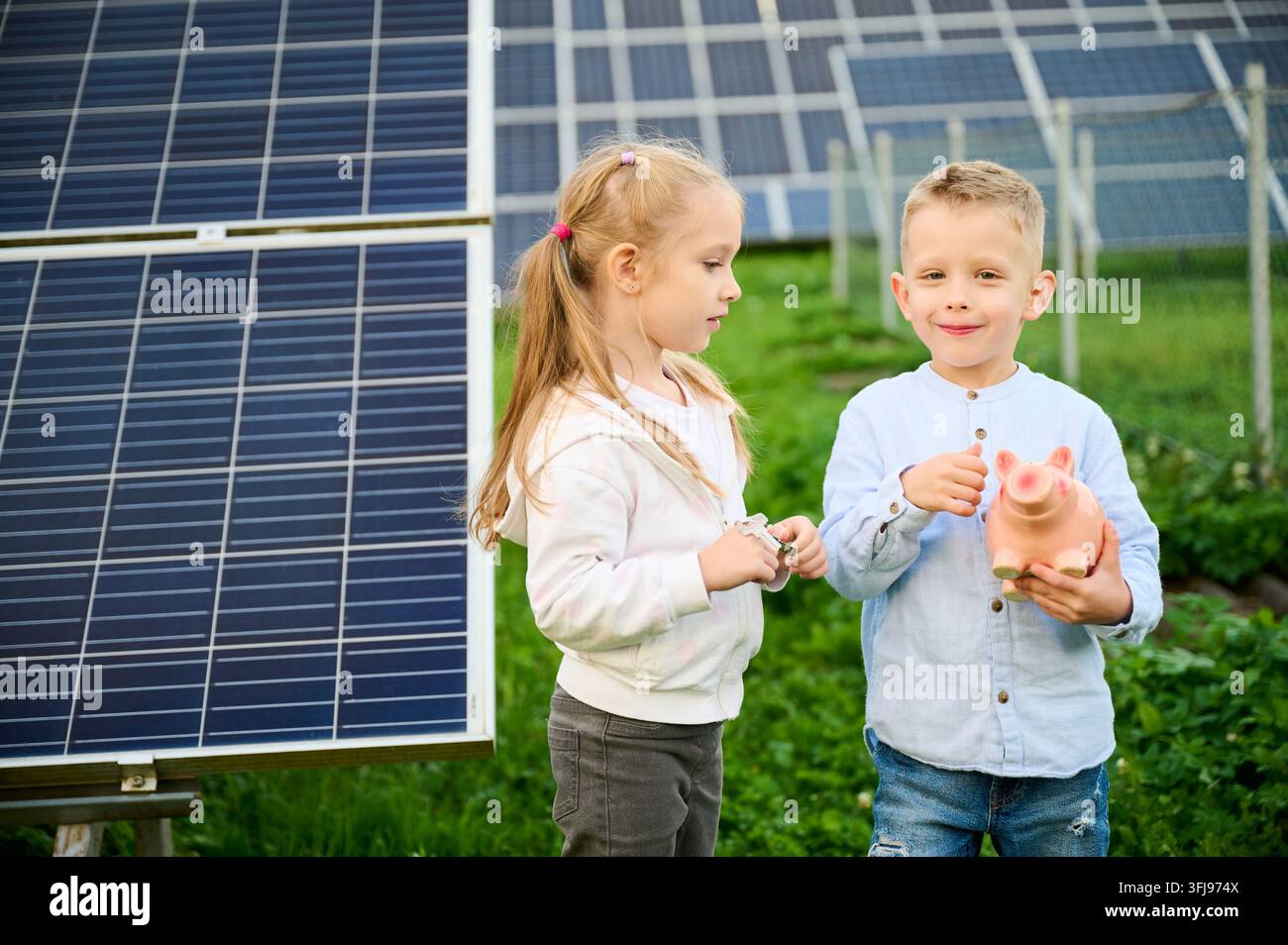 Ragazzo che tiene un salvadanaio e sorride mentre una ragazza tiene una moneta vicino ai pannelli solari. Bambini che risparmiano denaro e imparano le energie rinnovabili. Concetto di finanza, sostenibilità, tecnologia pulita Foto Stock