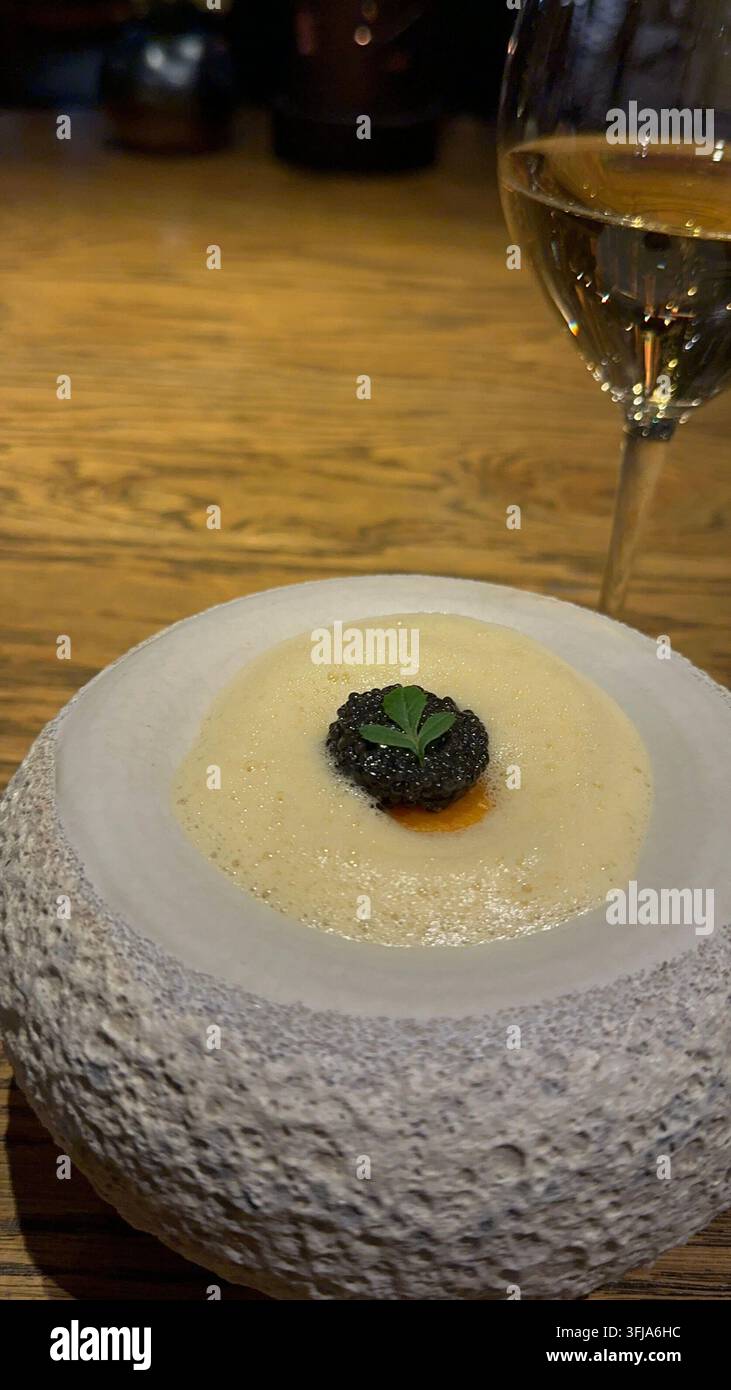 Piatto gourmet guarnito con caviale nero e un bicchiere di vino bianco. Piatto gourmet servito in una ciotola di pietra strutturata, con una salsa cremosa ricoperta di blè Foto Stock