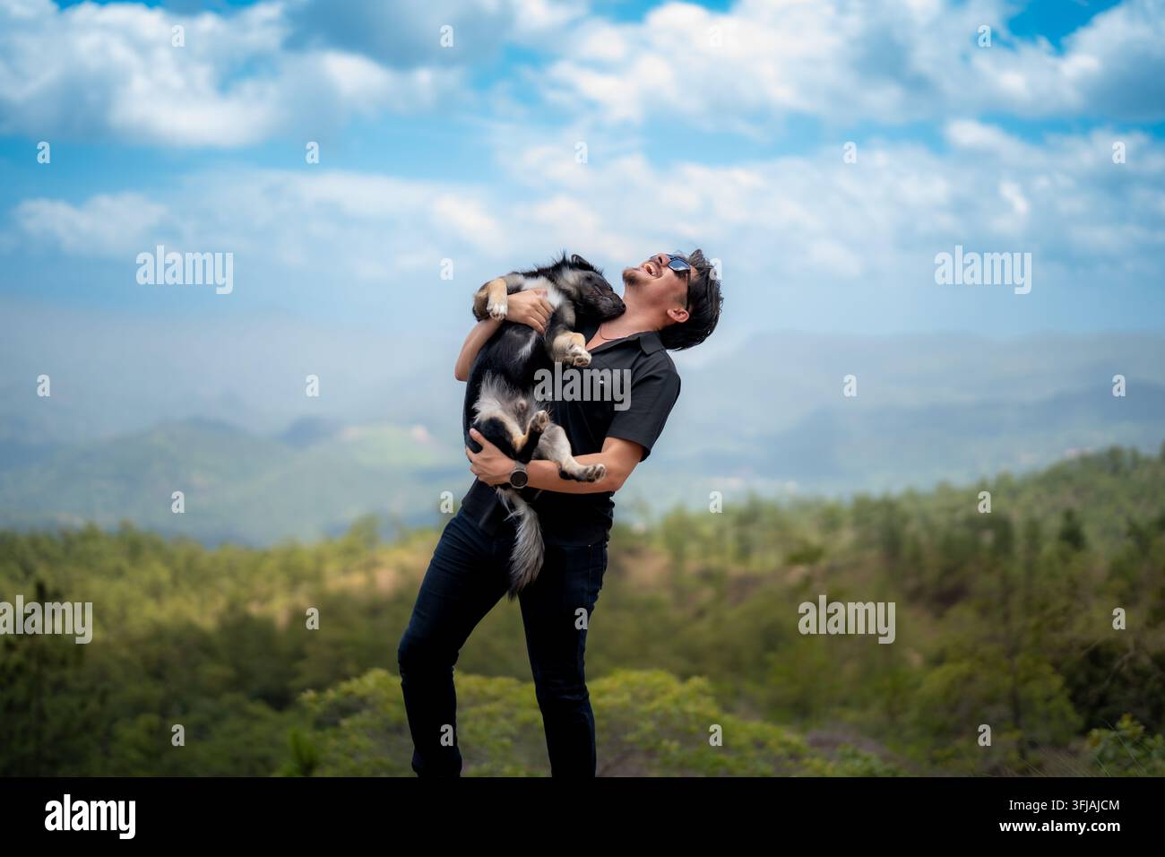 Giovane uomo gioioso in camicia nera e occhiali da sole che ride mentre abbraccia il suo cane sulla cima di una montagna Foto Stock