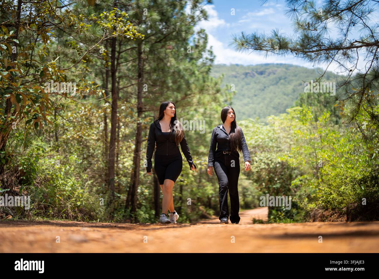 Due giovani donne in abiti neri che camminano in avanti su una strada sterrata rossa in una foresta di pini Foto Stock