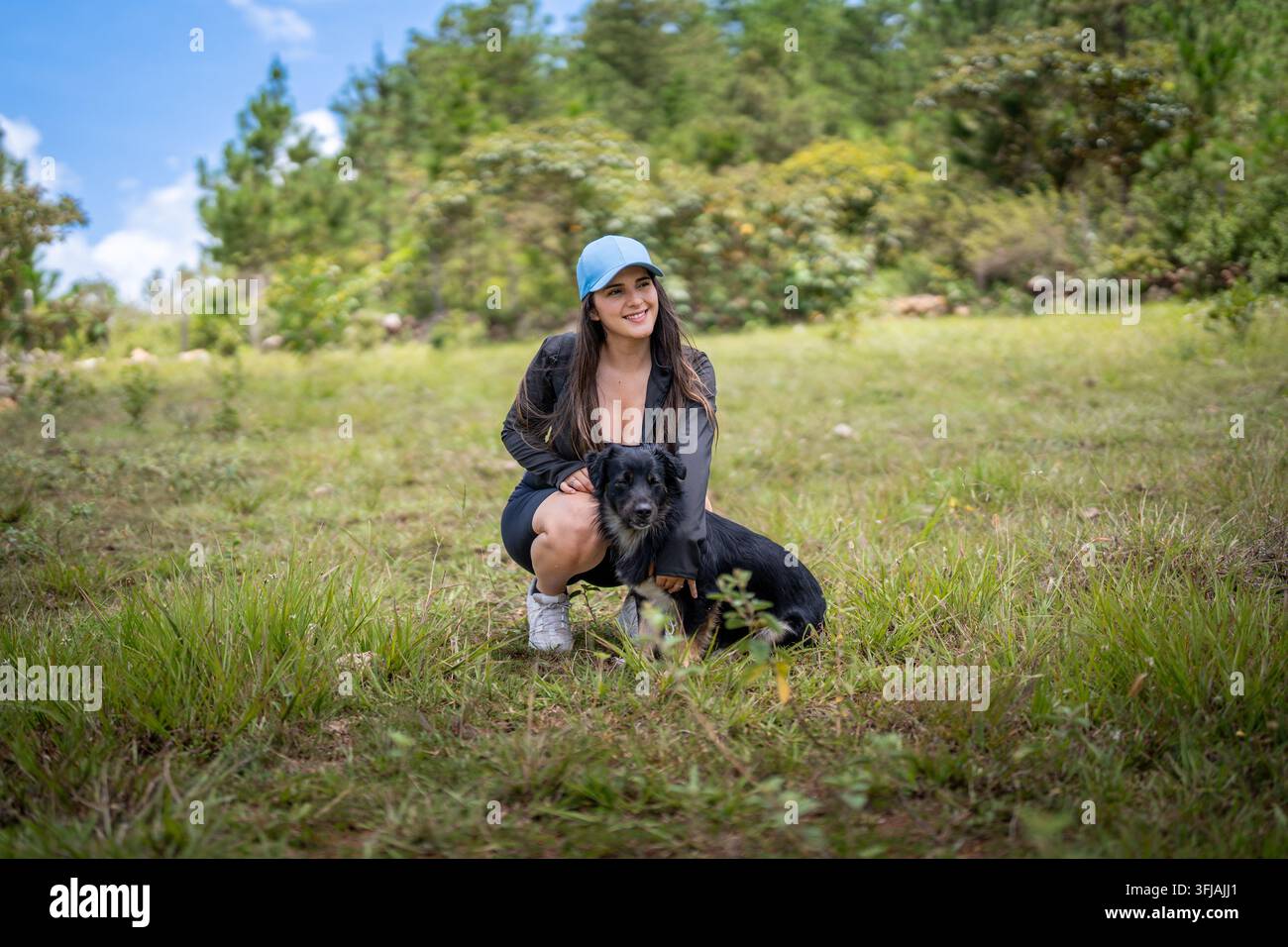 Giovane donna sorridente con cappellino sportivo blu, accovacciata e che abbraccia il suo cane in un prato verde Foto Stock