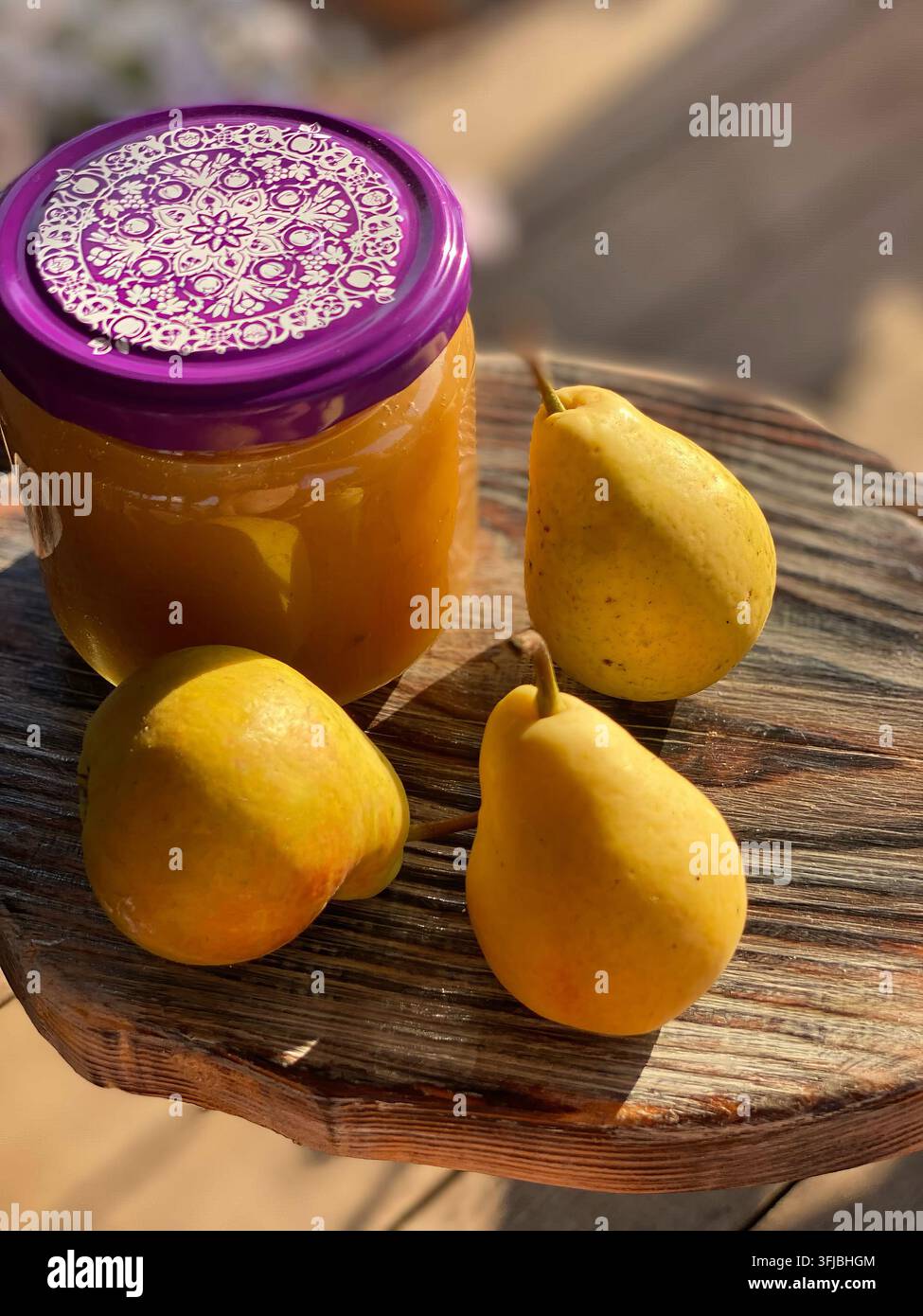 Marmellata di pere dorate in vasetto di vetro accanto a tre pere gialle fresche su legno stagionato. La luce calda crea ombre morbide che evidenziano la texture lucida Foto Stock