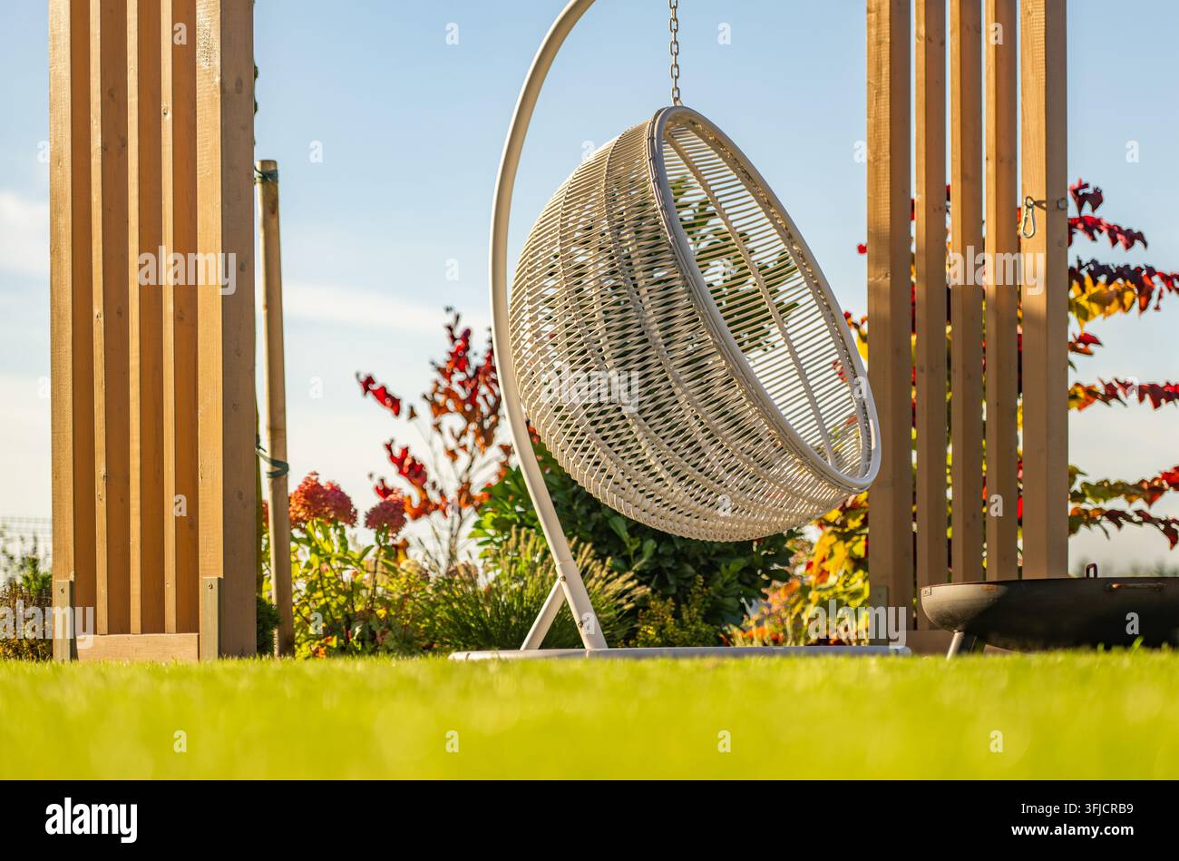 Una sedia sospesa tessuta vuota è appesa a una struttura metallica autoportante tra i pali di una pergola in legno. Un prato e un bordo del giardino piantato circondano il Foto Stock