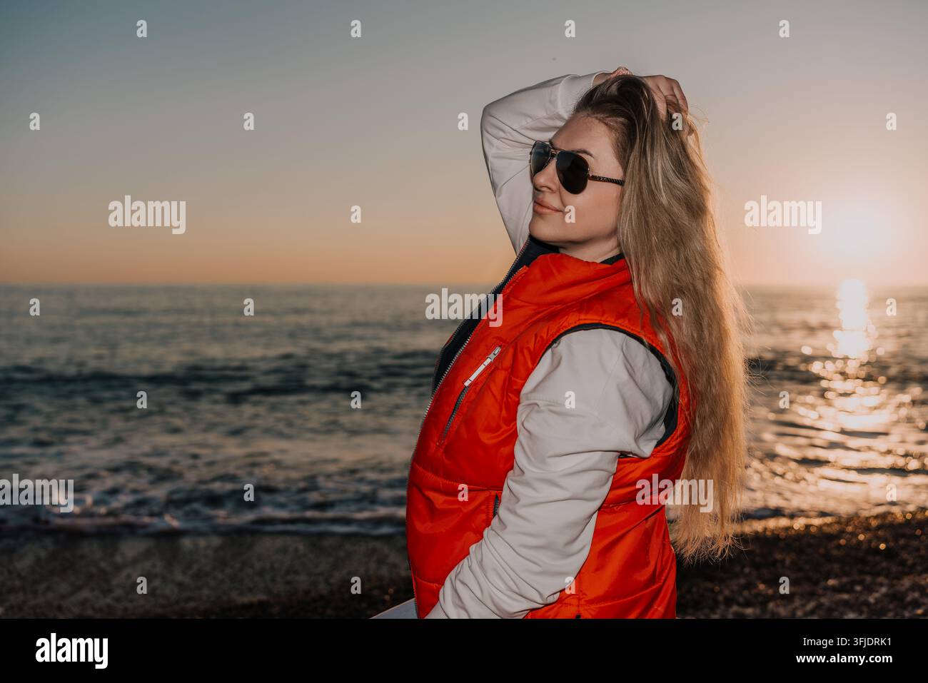 Donna in posa sulla spiaggia al tramonto con giacca a piumino rossa durante l'ora dorata con vista sull'oceano spazio di copia Foto Stock