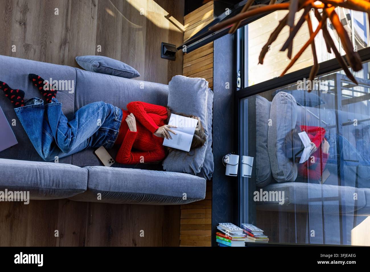 Giovane donna che copre il viso con un libro sul divano. Giovane donna in maglione rosso sdraiata su un divano grigio con un libro aperto a faccia in giù che le copre il viso. Foto Stock