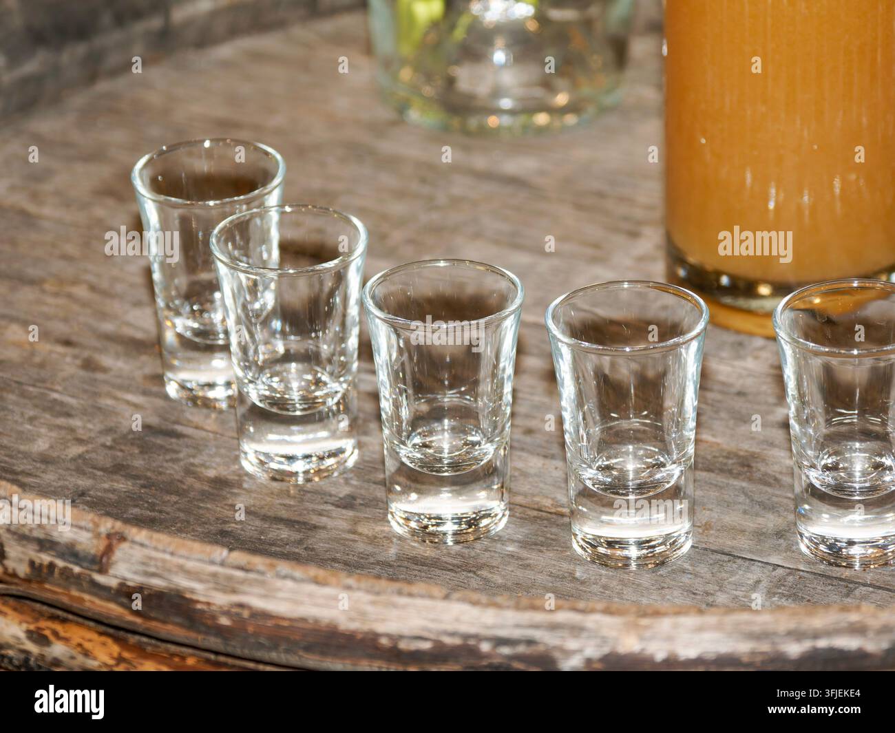 Cinque bicchieri da shot vuoti disposti in fila su una superficie di legno rustico con una bevanda parzialmente visibile in un contenitore. Foto Stock