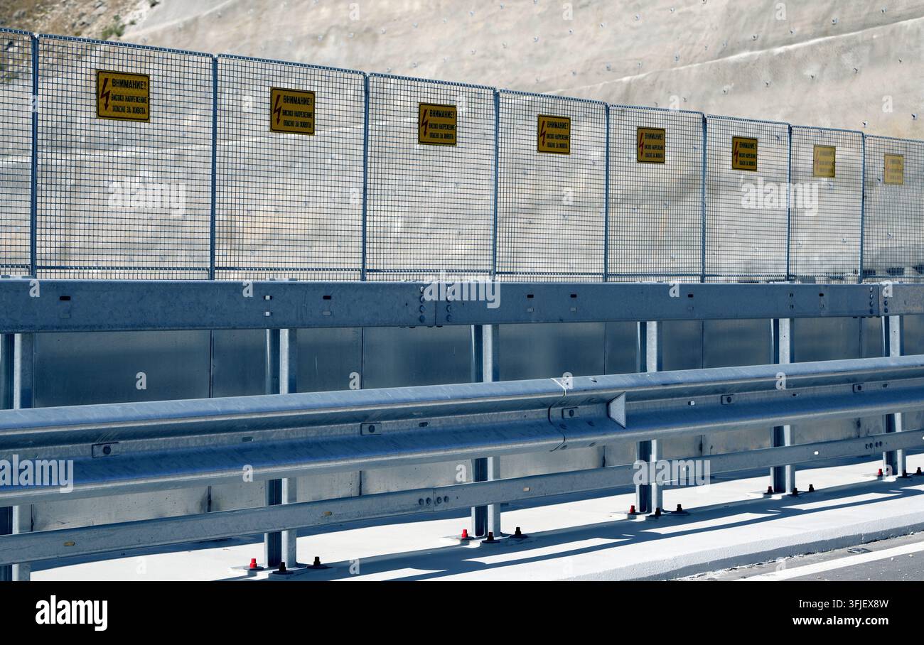 Guardrail in acciaio e recinzione in rete metallica che proteggono una strada, con più segnali di avvertimento gialli che indicano pericolo di alta tensione, provi Foto Stock