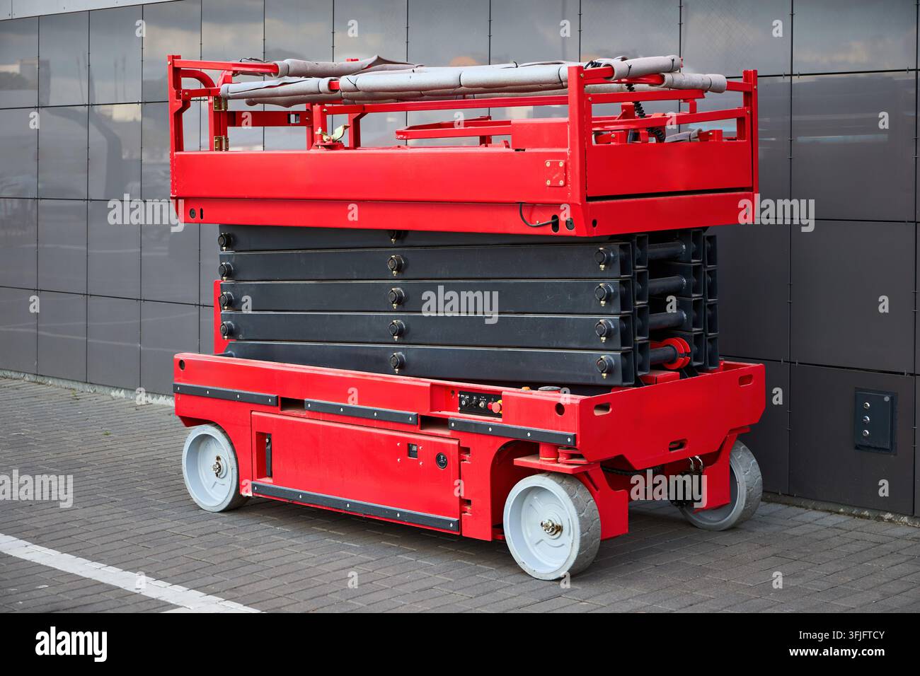 Piattaforma elevatrice a forbice rossa parcheggiata all'aperto vicino al muro di un edificio moderno, attrezzatura di sollevamento idraulica industriale, piattaforma di lavoro mobile usata per Con Foto Stock