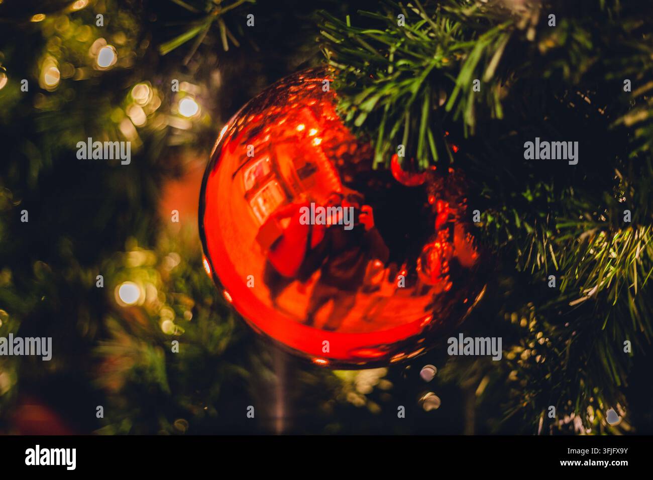 Prima piano di una sfera natalizia rossa lucida appesa a un albero decorato, che riflette un fotografo con una fotocamera all'interno. Foto Stock