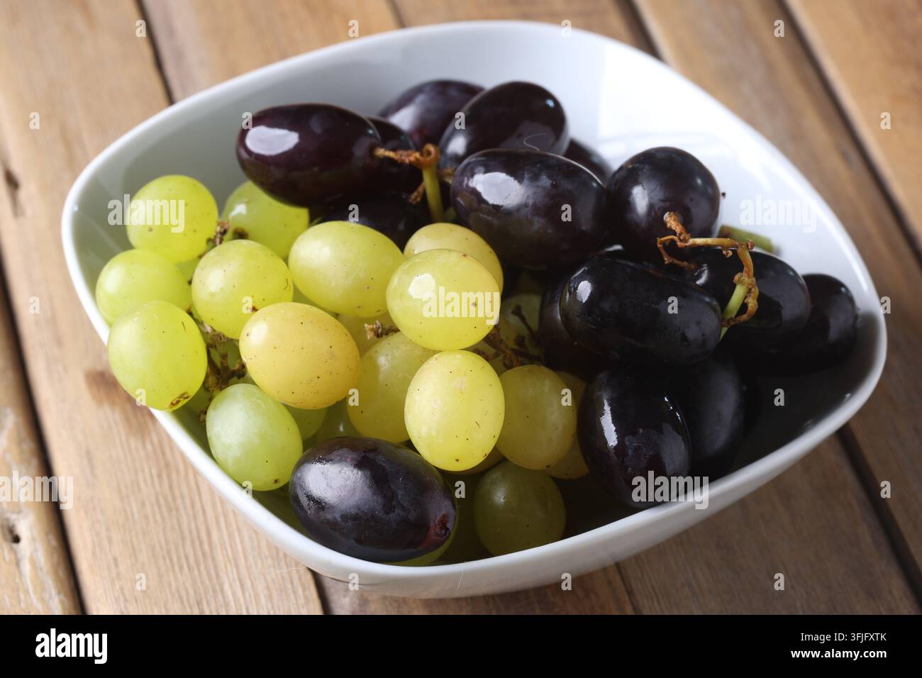 Uva verde e rossa fresca disposta in una ciotola bianca su un tavolo di legno Foto Stock