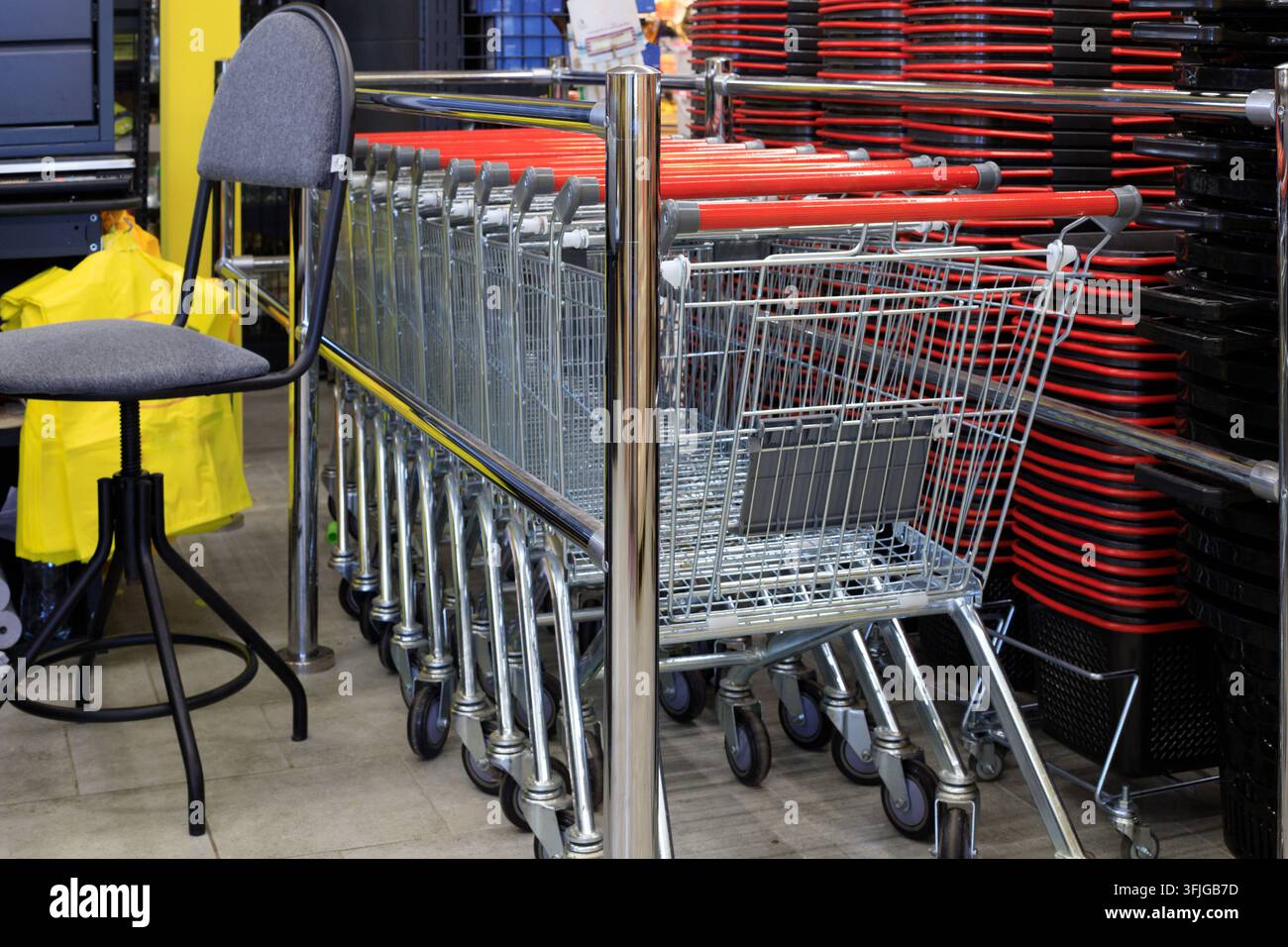 Carrelli della spesa in metallo con manici rossi allineati vicino a ringhiere cromate accanto a cestini di plastica impilati e una sedia all'interno di un negozio al dettaglio. Foto Stock