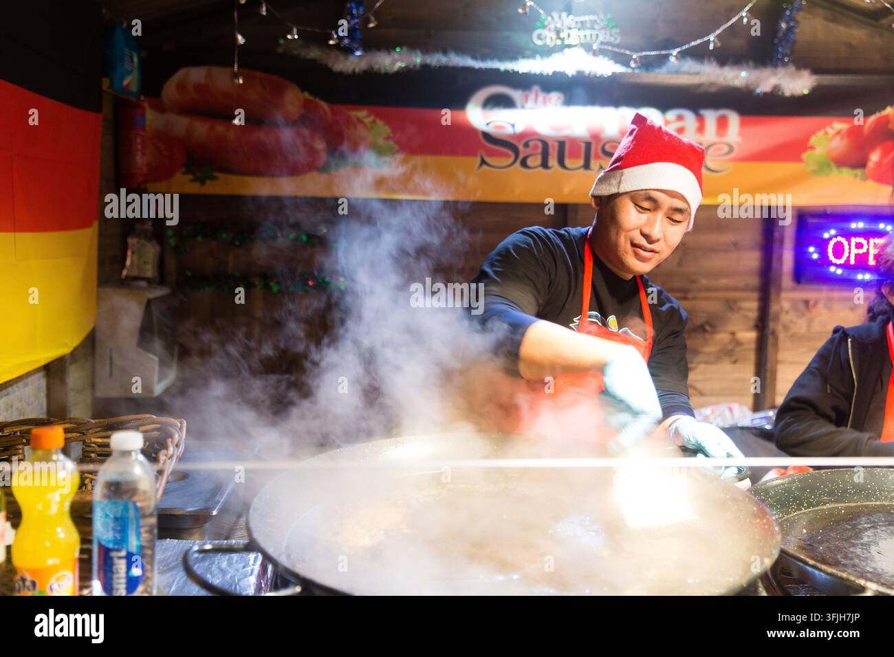 Un venditore con un cappello di Babbo Natale cucina salsicce tedesche al mercato natalizio di More London vicino al Tower Bridge per sfamare i visitatori delle festività. Foto Stock