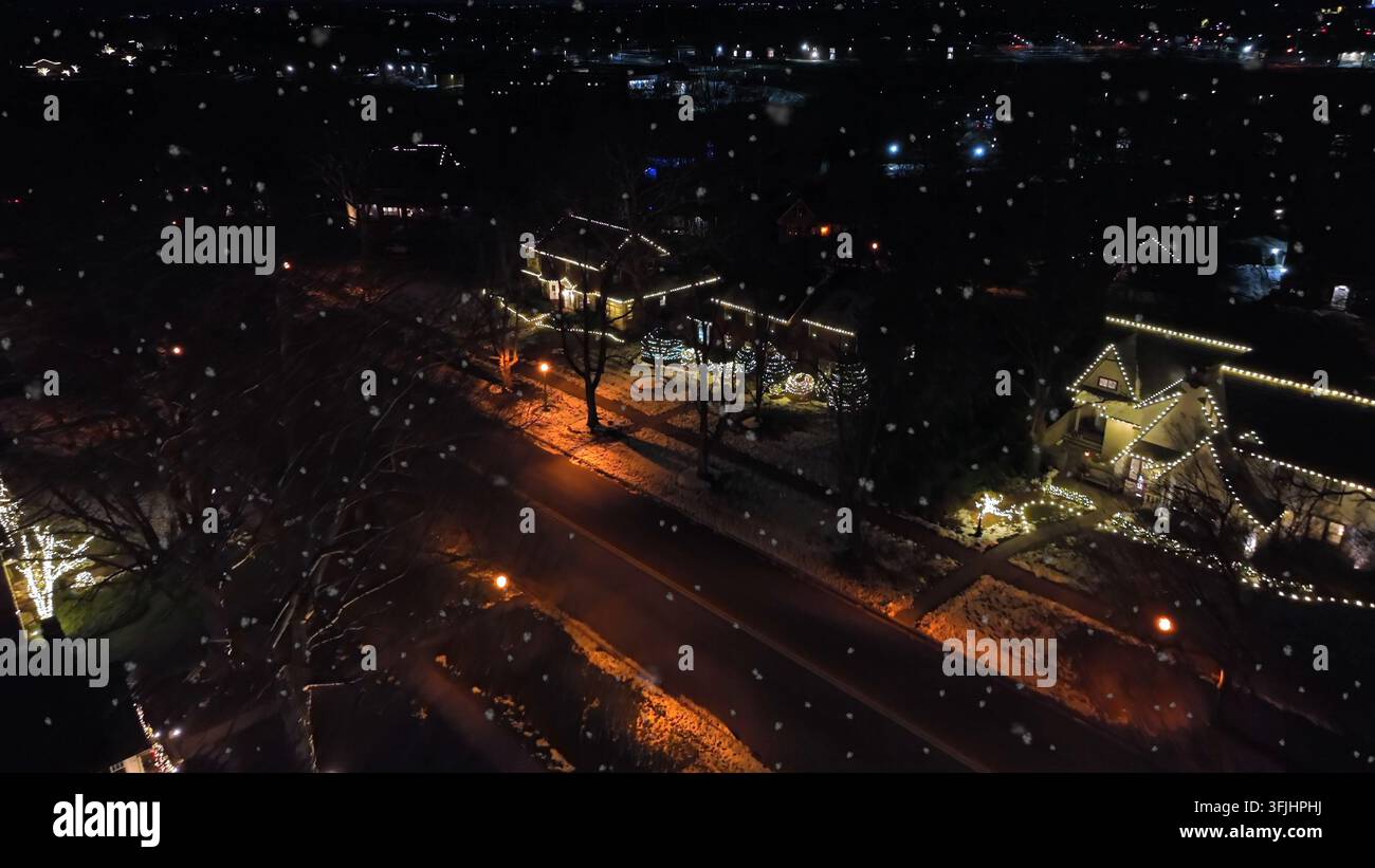 Vista aerea notturna di un quartiere suburbano innevato in America durante il Natale, decorazioni e luci luminose che illuminano le case mentre le auto si muovono lentamente Foto Stock