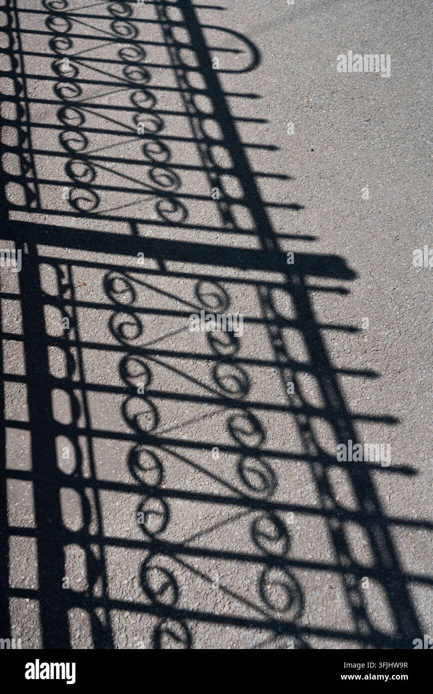 La luce del sole proietta l'ombra di un cancello metallico ornato sull'asfalto, creando schemi geometrici sorprendenti. Foto Stock