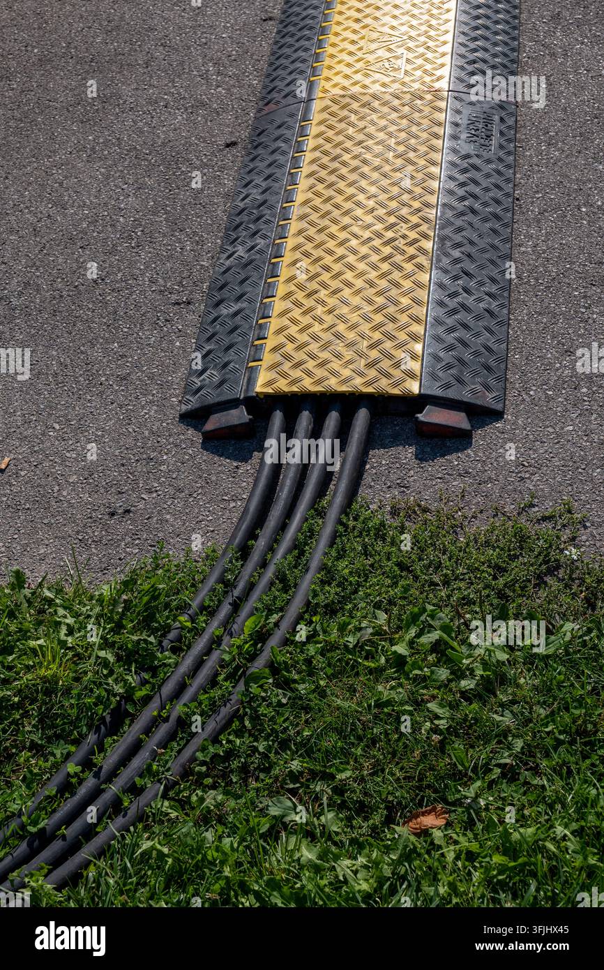 Rampa di protezione dei cavi gialla copre condotti neri che attraversano l'asfalto accanto all'erba. Foto Stock