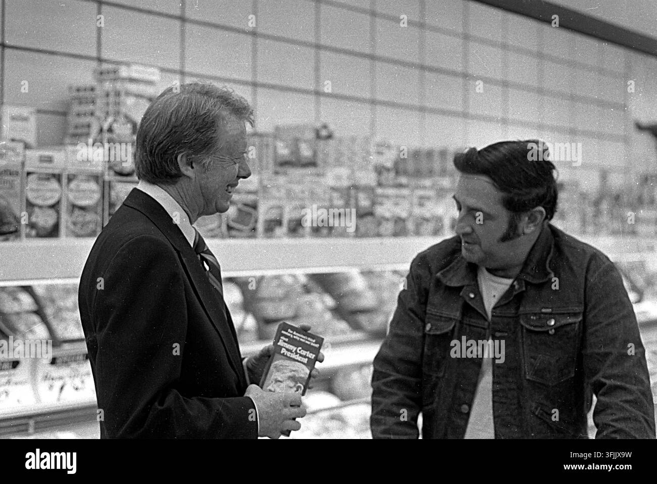Jimmy Carter fa campagna in un negozio di alimentari del New Hampshire il 18 aprile 1975. Sorride mentre tiene un volantino di campagna per un uomo con una giacca di jeans. Foto Stock