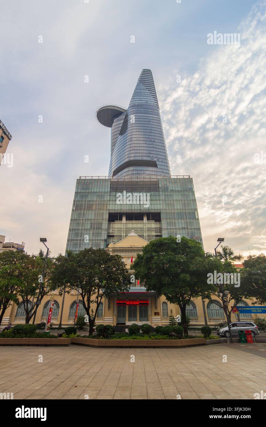 Un grattacielo di vetro curvo si erge dietro un edificio civico storico, alberi e un'ampia piazza lastricata che mostra striscioni politici vietnamiti. Foto Stock