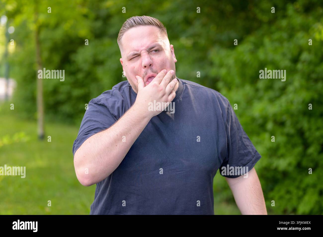 Giovane uomo stanco che sbadiglia mentre si copre la bocca all'aperto in un ambiente naturale verdeggiante, esprimendo stanchezza, esaurimento, sonnolenza, burnout e mancanza Foto Stock