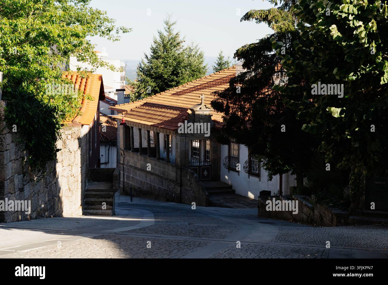 Guarda, Portogallo – 12 settembre 2026. Una strada di ciottoli vuota scende accanto a una casa in granito con tetto in terracotta, croce di pietra e galleria colonnata Foto Stock