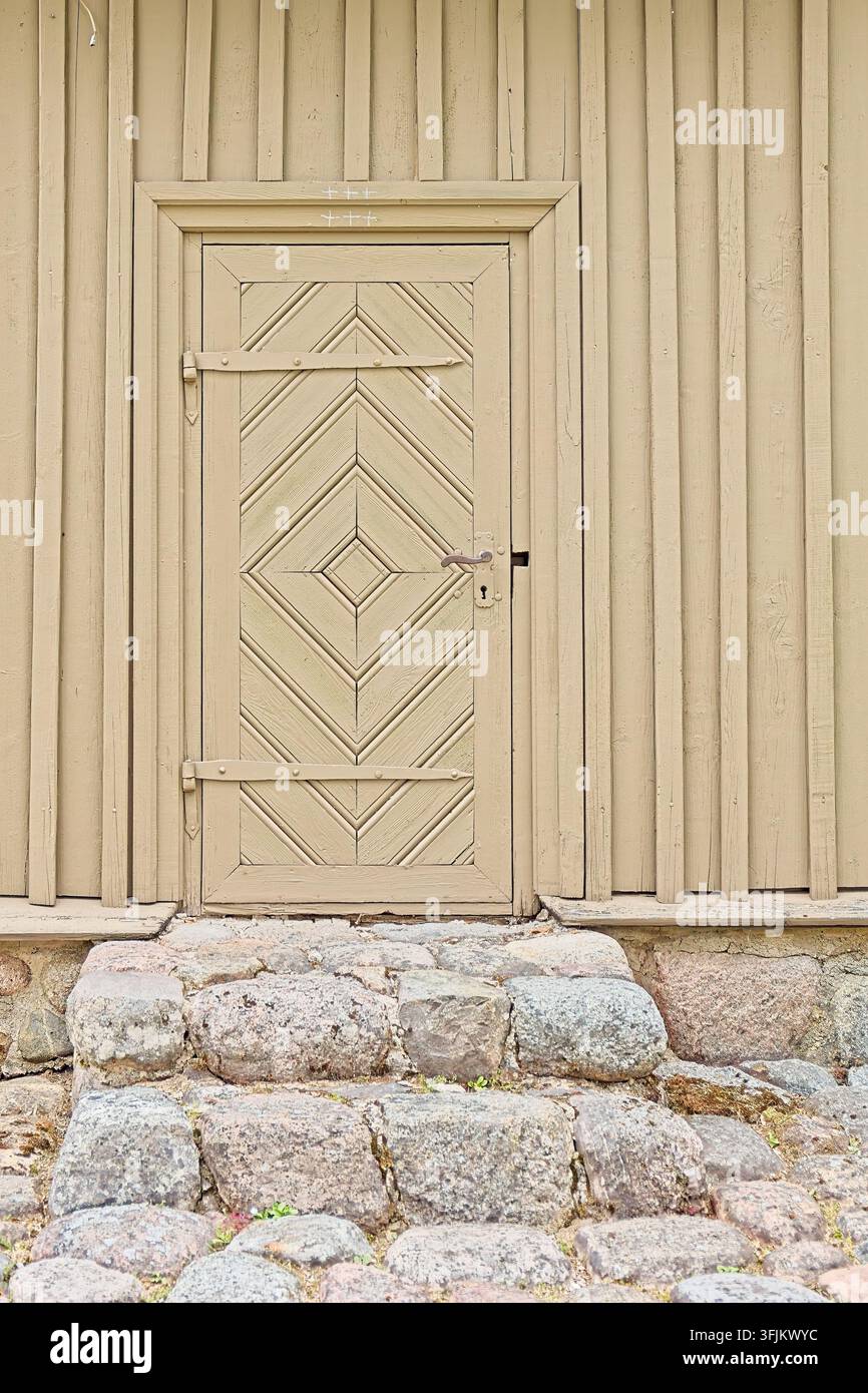 Antica porta di legno con un motivo geometrico a rombo, lunghe cerniere di ferro e una maniglia semplice, inserita in un muro di legno color sabbia chiaro, Lituania. Foto Stock
