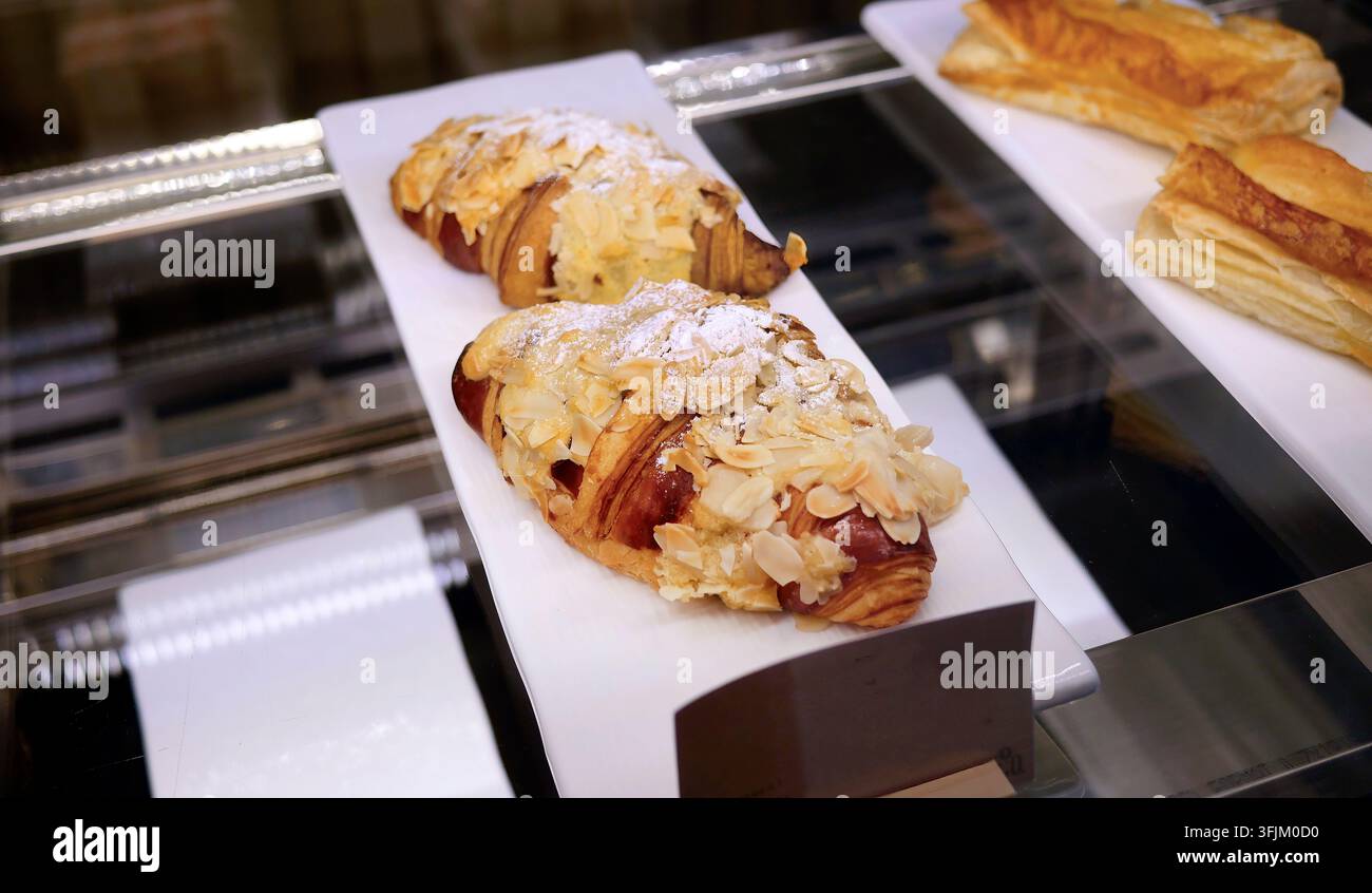 Close up di croissant alle mandorle appena sfornati su piatto rettangolare bianco all'interno di una vetrina da forno in vetro. Pasticceria francese tradizionale ricoperta di aglio affettato Foto Stock