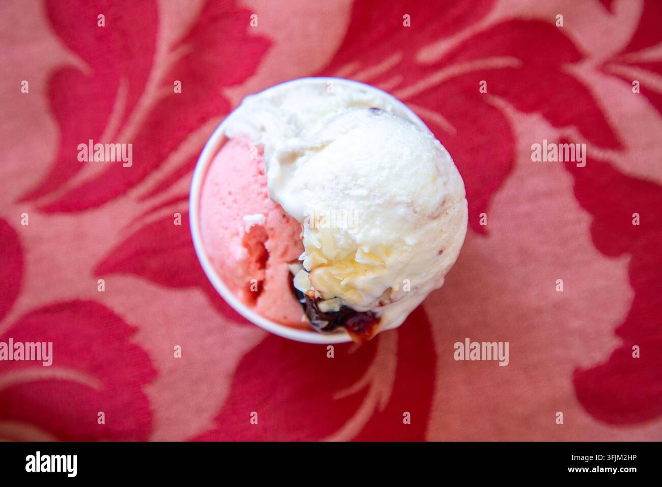 Vista dall'alto di due cucchiaiate di gelato rosa e bianco in una piccola ciotola su un tessuto floreale rosso vivace con sfocatura morbida, texture cremosa e giocosa estate Foto Stock