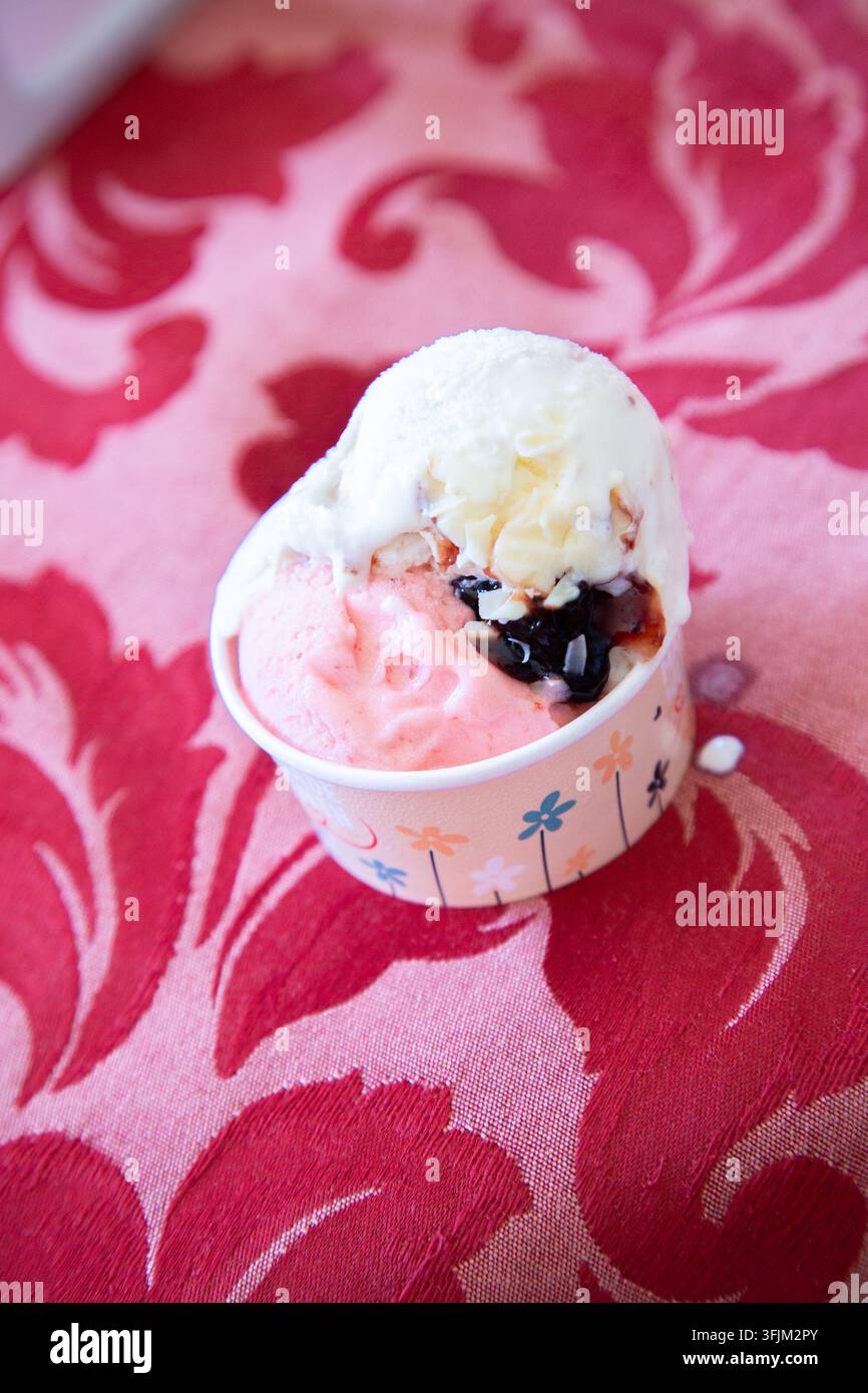 Copa di gelato colorata con palline rosa e bianche ricoperte di salsa scura, appoggiata su un tessuto floreale rosso riccamente strutturato alla luce naturale soffusa, creando Foto Stock