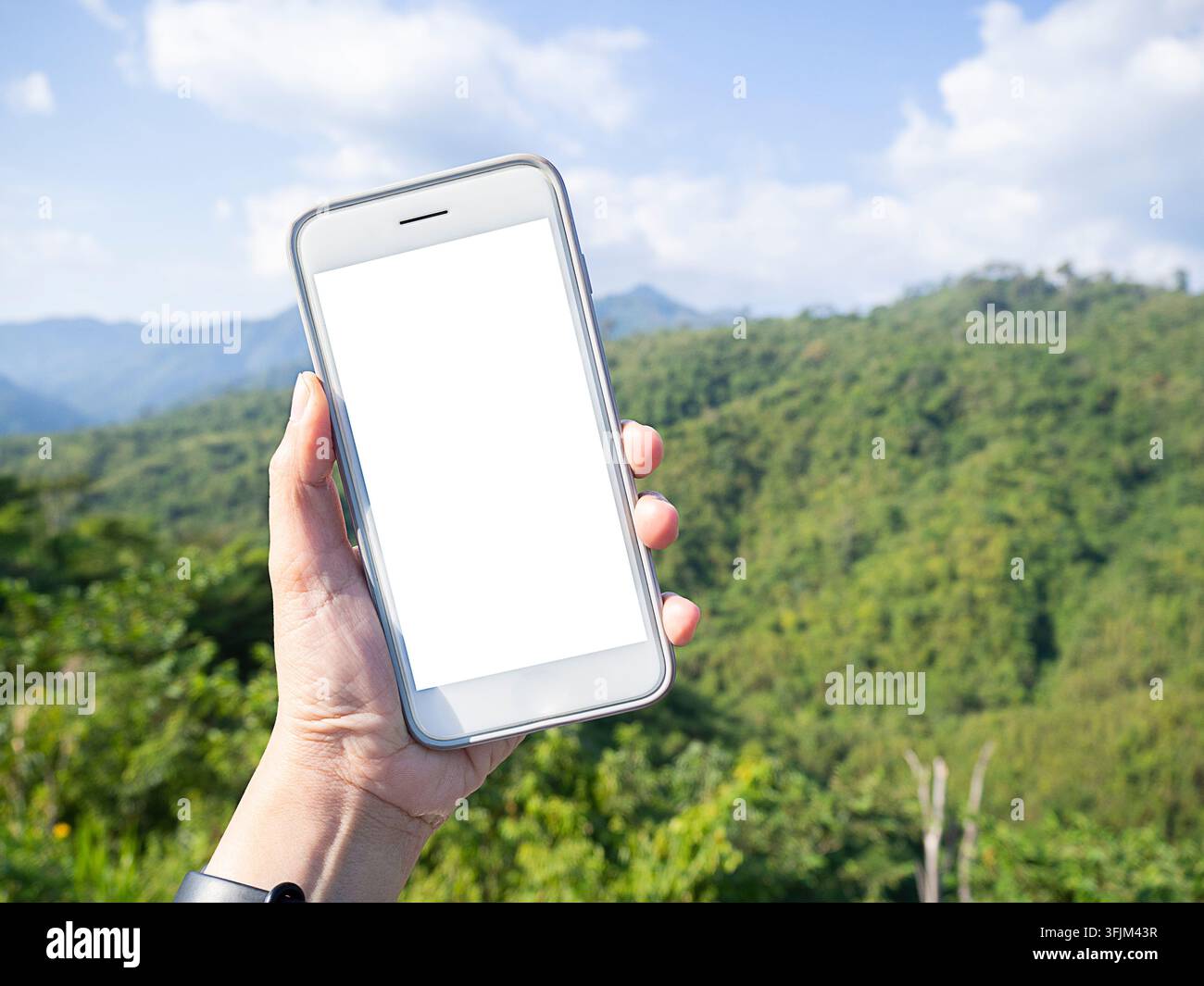 Immagine mockup di una mano che tiene e mostra uno smartphone bianco con schermo desktop vuoto all'aperto con sfondo sfocato di montagne verdi. Foto Stock